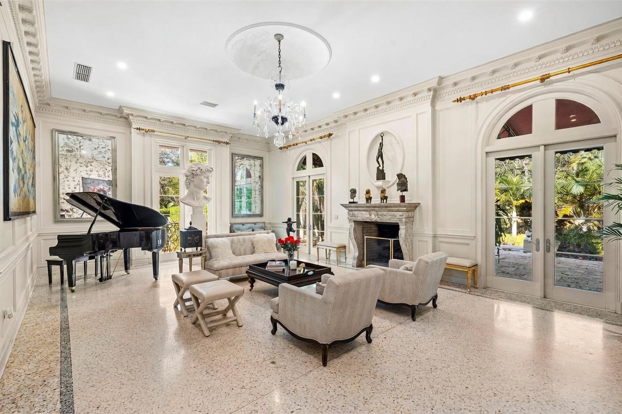 This elegant, formal living room features high ceilings with intricate crown molding and a central crystal chandelier. The space is anchored by a stone fireplace with a decorative mantel and is furnished with a classic sofa, armchairs, and a grand piano. Large glass doors provide abundant natural light and views of the lush outdoor greenery, creating a sophisticated and airy atmosphere.