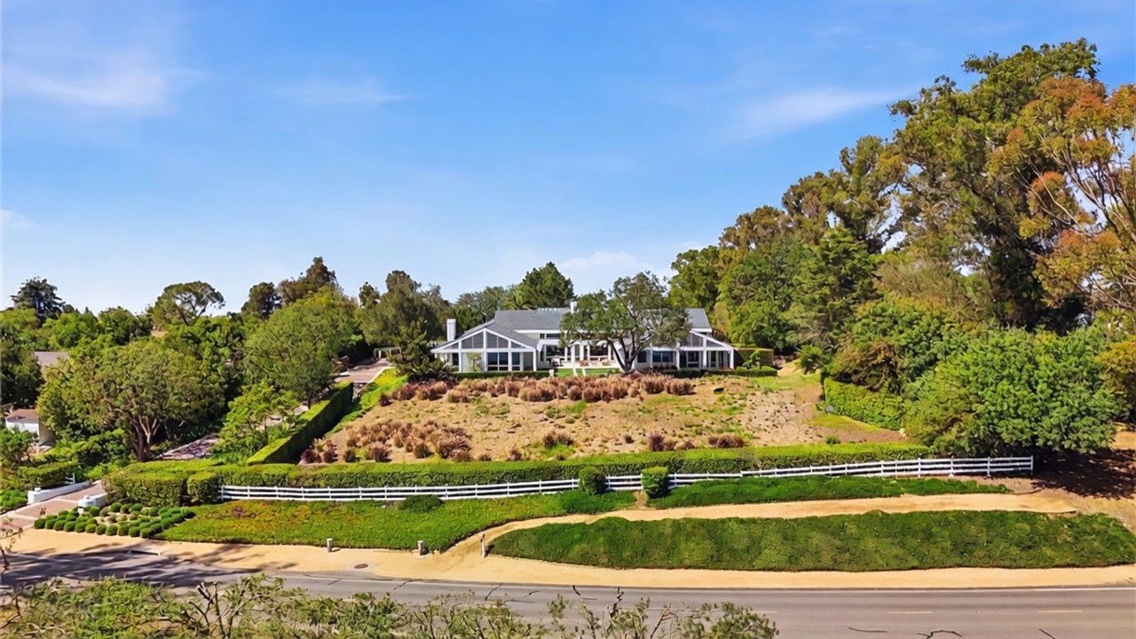 This elevated perspective captures a sprawling single-story residence set back from a quiet road on a large, landscaped lot. The home features a classic architectural style with a multi-gabled roofline, expansive glass windows, and a neutral exterior finish. A white picket fence borders the property, separating the manicured lawn and terraced hillside from the street below, creating a sense of privacy and suburban charm.