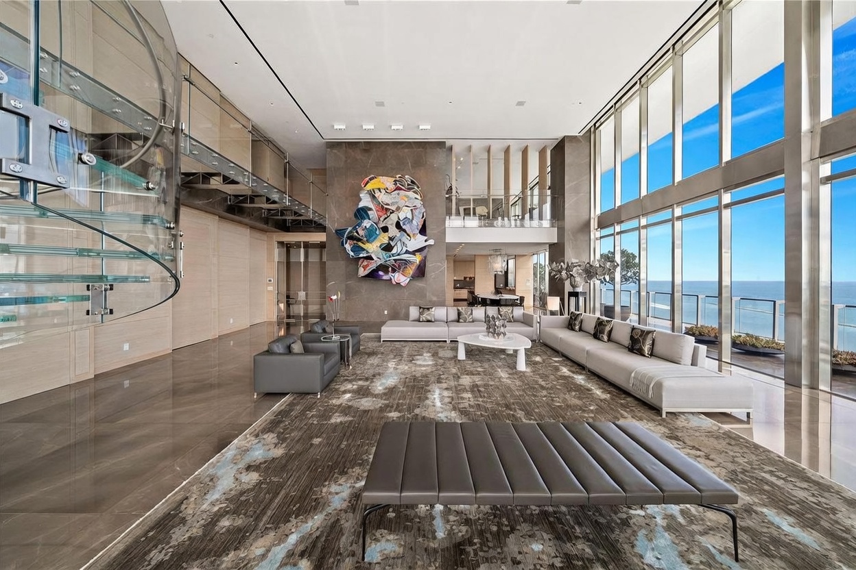 This expansive, double-height living room features a dramatic glass spiral staircase and a large, abstract wall sculpture mounted on a dark stone feature wall. The space is defined by floor-to-ceiling windows that offer panoramic ocean views, complemented by a neutral color palette and a large, textured area rug. The open-concept layout creates an airy, sophisticated atmosphere, perfect for luxury entertaining.