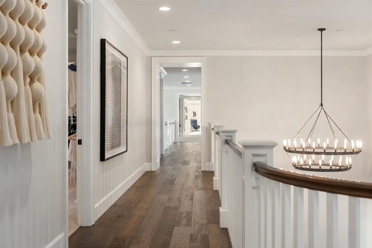 This bright, elegant hallway features rich, dark wood flooring and crisp white walls, creating a sophisticated transition between living spaces. A large, multi-tiered circular chandelier hangs over the stairwell, while a textured wall hanging and a framed geometric print add artistic character to the corridor. The perspective looks down the length of the hallway, showcasing a series of doorways that lead into further rooms, emphasizing the home's open and airy layout.