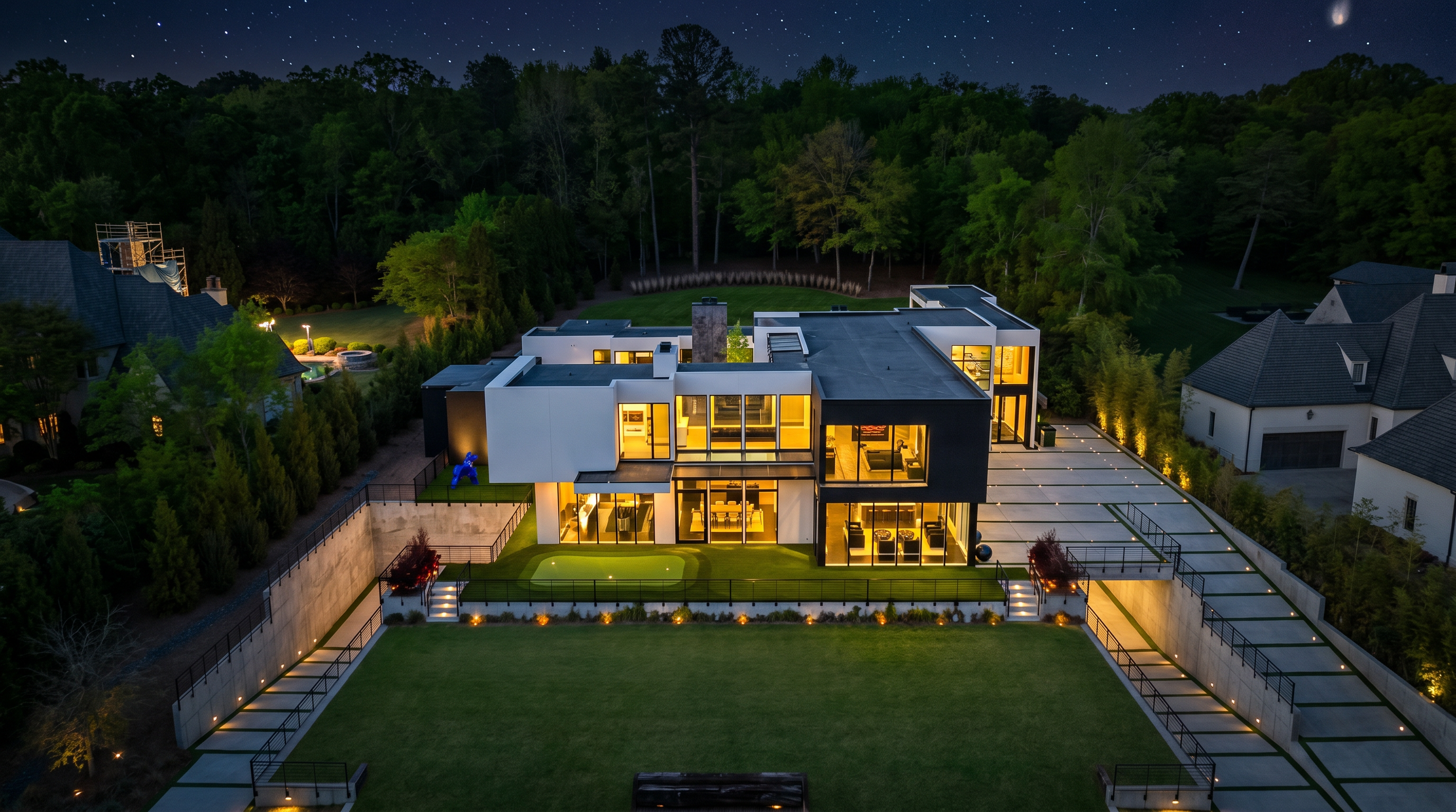 This striking aerial view captures a modern, multi-level luxury residence characterized by its bold geometric architecture and contrasting black and white exterior finishes. The property features an expansive backyard with a manicured lawn, a putting green, and a tiered retaining wall, all set against a lush backdrop of mature trees under a dramatic sunset sky. The home's design emphasizes clean lines, large windows, and a sophisticated layout that seamlessly integrates indoor and outdoor living spaces.