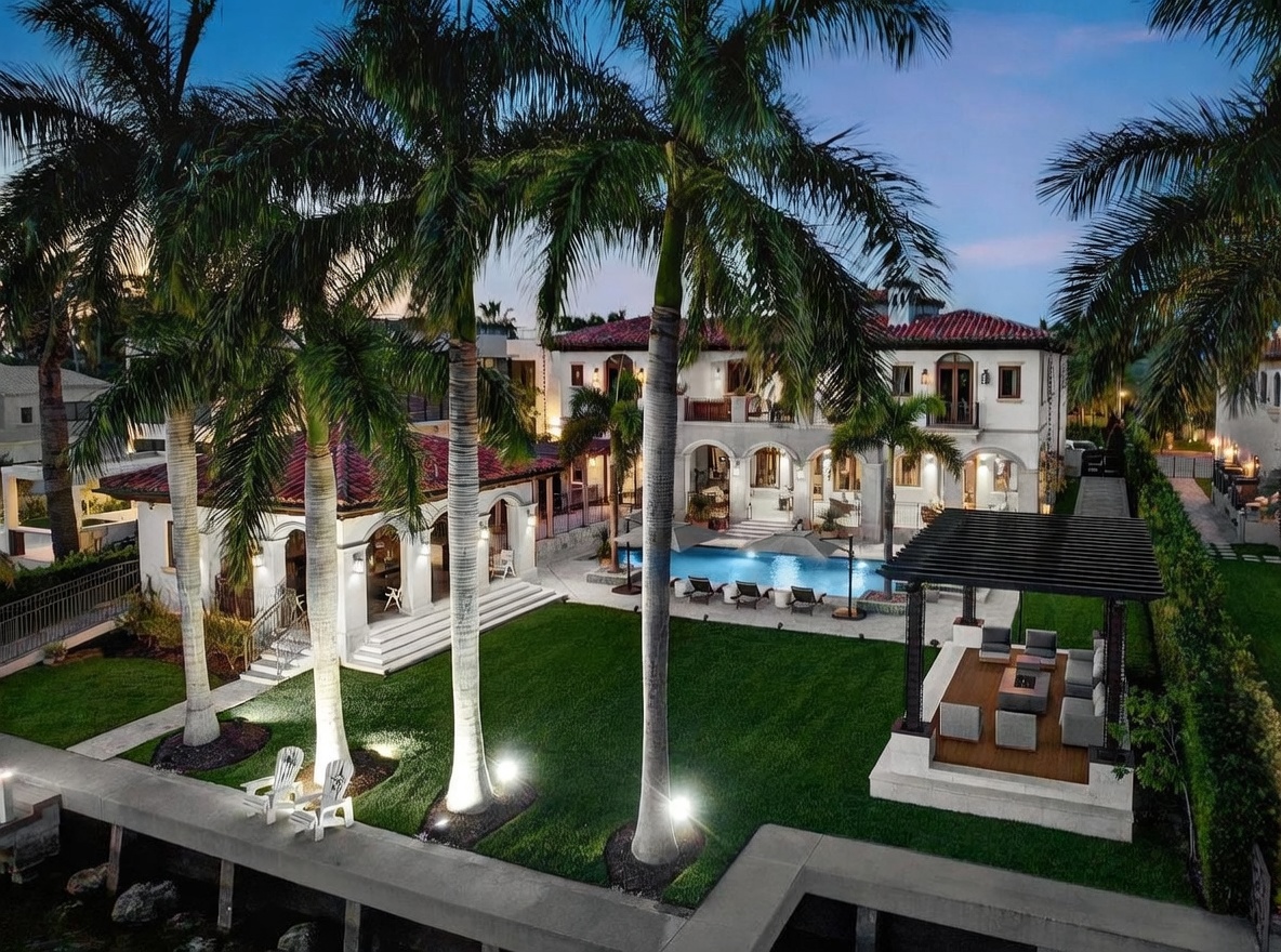 This stunning aerial view captures a luxurious waterfront estate at dusk, featuring a Mediterranean-style mansion with a red-tiled roof and expansive outdoor living spaces. The property boasts a pristine swimming pool, a manicured lawn lined with tall palm trees, and a covered pergola with a fire pit area overlooking the water. The warm ambient lighting creates an inviting and sophisticated atmosphere, highlighting the high-end architectural details and resort-like amenities.