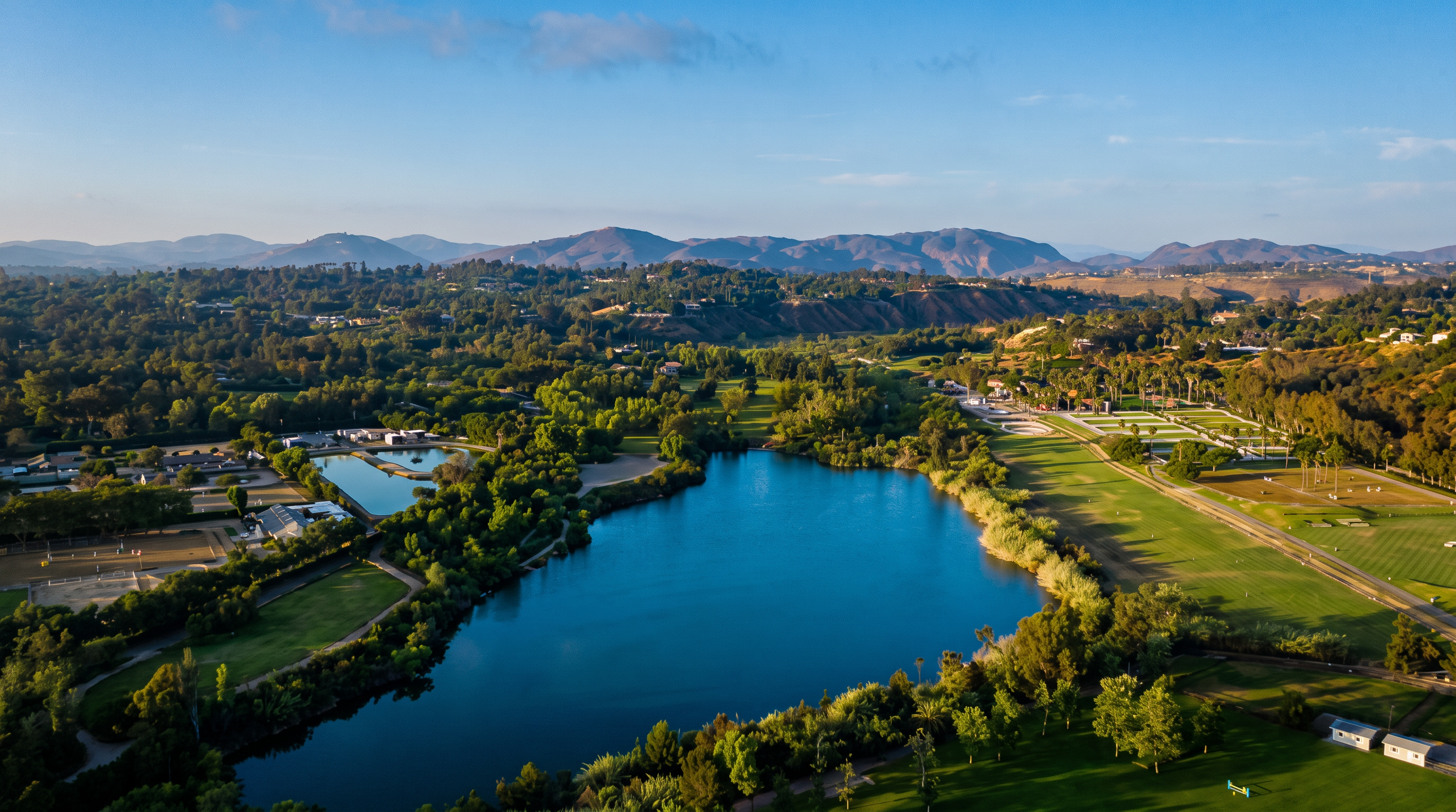 This high-angle aerial view captures a sprawling landscape featuring a large, central lake surrounded by lush greenery and mature trees. To the right, a manicured golf course stretches out with visible fairways and sand traps, while the background showcases rolling hills under a clear blue sky. The perspective provides a comprehensive look at the expansive outdoor recreational space and the natural beauty of the surrounding environment.