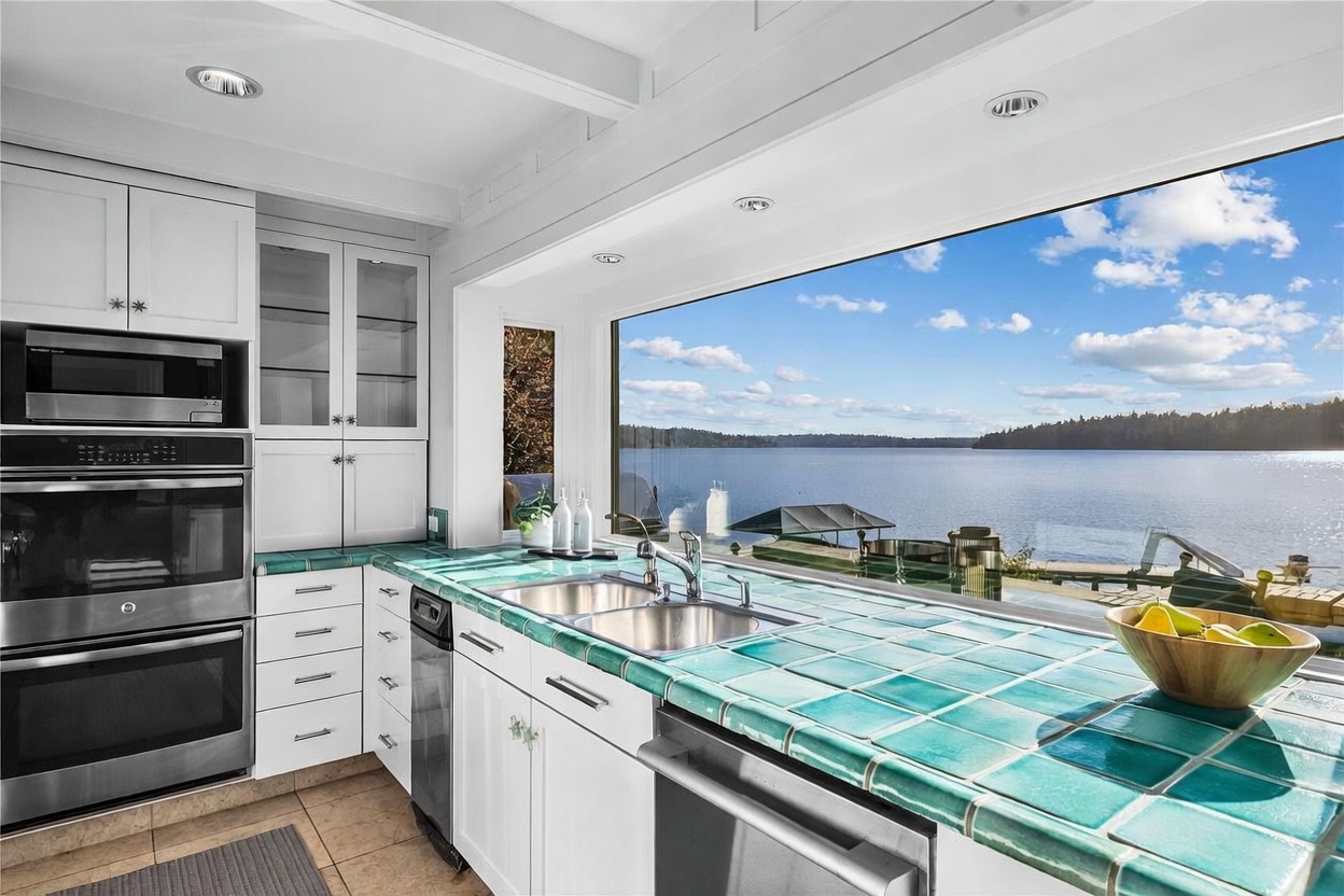 This bright kitchen features white cabinetry paired with vibrant teal tiled countertops, creating a unique, coastal-inspired aesthetic. A large, expansive window serves as the focal point, offering a stunning, unobstructed panoramic view of the lake and surrounding landscape. The space is equipped with built-in stainless steel appliances, including a double oven and microwave, blending functional utility with a serene, scenic atmosphere.