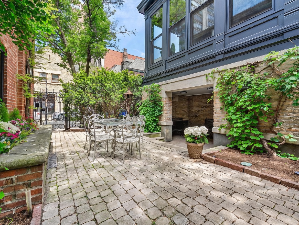This charming brick-paved patio offers a private outdoor retreat, featuring a wrought-iron dining set and a covered alcove built into the home's foundation. Lush greenery, including climbing vines and potted hydrangeas, softens the architectural lines of the brick and dark-painted exterior walls. The perspective captures a serene, enclosed courtyard atmosphere, perfect for quiet relaxation or intimate outdoor dining.