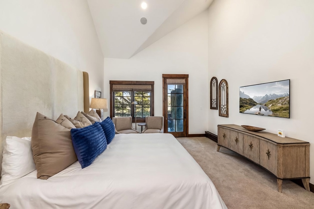 This spacious primary bedroom features a high vaulted ceiling and a large, light-colored upholstered headboard that serves as a focal point. The room includes a comfortable seating area near the windows, a wooden media console beneath a wall-mounted television, and decorative arched wall accents. The neutral color palette and warm wood trim create a serene and inviting atmosphere.