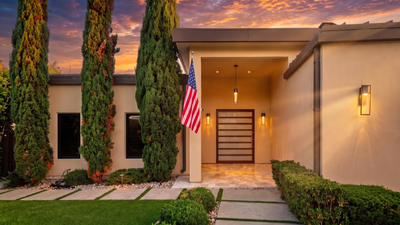 This modern home features a striking entryway with a horizontal-slatted wooden door and a recessed porch area. The exterior is accented by tall, manicured cypress trees and a clean, geometric walkway made of concrete pavers set into a gravel and grass landscape. An American flag hangs prominently near the entrance, adding a classic touch to the contemporary architectural design.