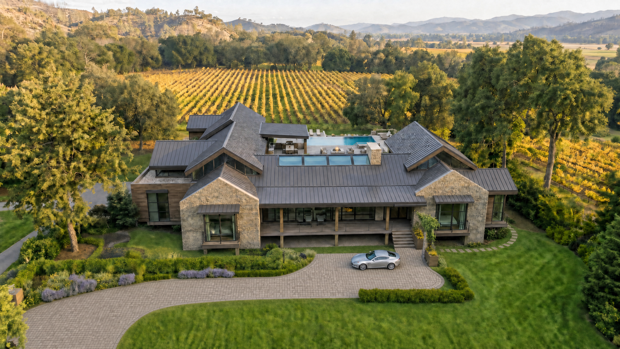 This stunning rural estate is captured from an elevated aerial perspective, showcasing its expansive property and sophisticated architectural design. The home features a complex, shingled roofline, stone cladding, and a sleek modern aesthetic, seamlessly integrated into a landscape of verdant lawns and lush vineyards. A paved driveway leads up to the home where a luxury sports car is parked, emphasizing the property's premium appeal and tranquil environment.