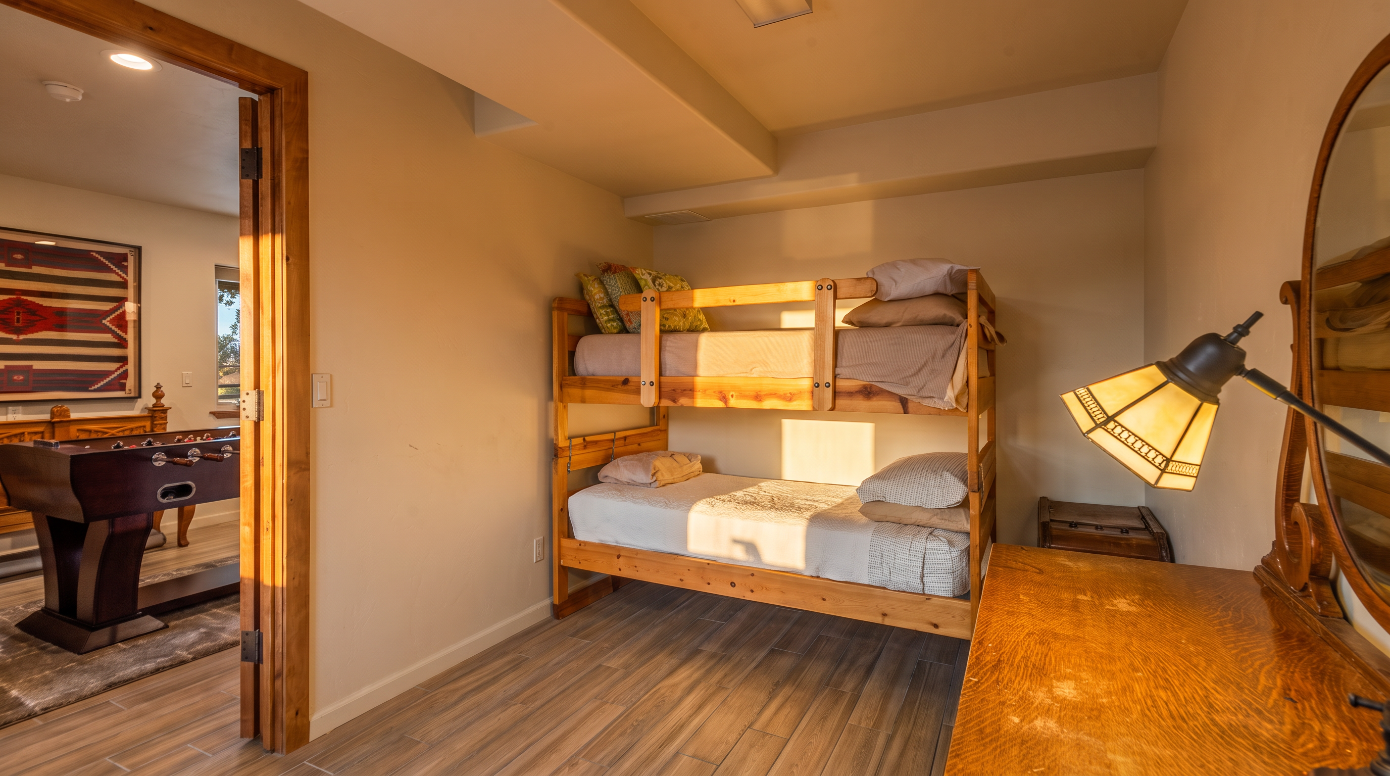 This guest bedroom features a sturdy wooden bunk bed with neutral bedding, providing a practical sleeping solution. The room is accented by a warm-toned wooden desk with a vintage-style lamp and leads into a games room housing a foosball table through an open wooden doorway. Wide-plank wood-look tile flooring flows throughout the space, creating a cohesive and rustic aesthetic.
