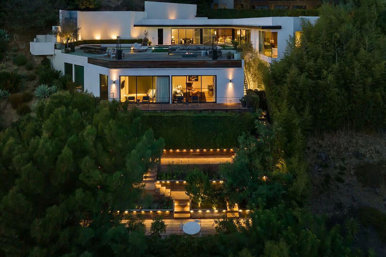 This aerial view captures a contemporary multi-level residence nestled into a hillside, illuminated at dusk. The home features a spacious upper terrace with a pool and lounge area, while the lower level includes a deck with large glass sliding doors that open to the interior living spaces. A series of landscaped, terraced garden levels with integrated lighting leads down the slope, creating a sophisticated and inviting outdoor environment.