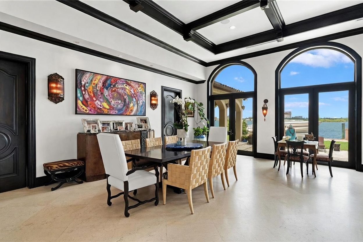 This elegant dining room features a dark wood table surrounded by a mix of upholstered and woven chairs, set against a backdrop of high ceilings with dark-toned coffered beams. Large arched windows and glass doors provide an expansive view of the waterfront and boat dock, flooding the space with natural light. The room is decorated with a vibrant abstract painting, wall-mounted sconces, and a sideboard displaying framed photos, creating a sophisticated and inviting atmosphere.