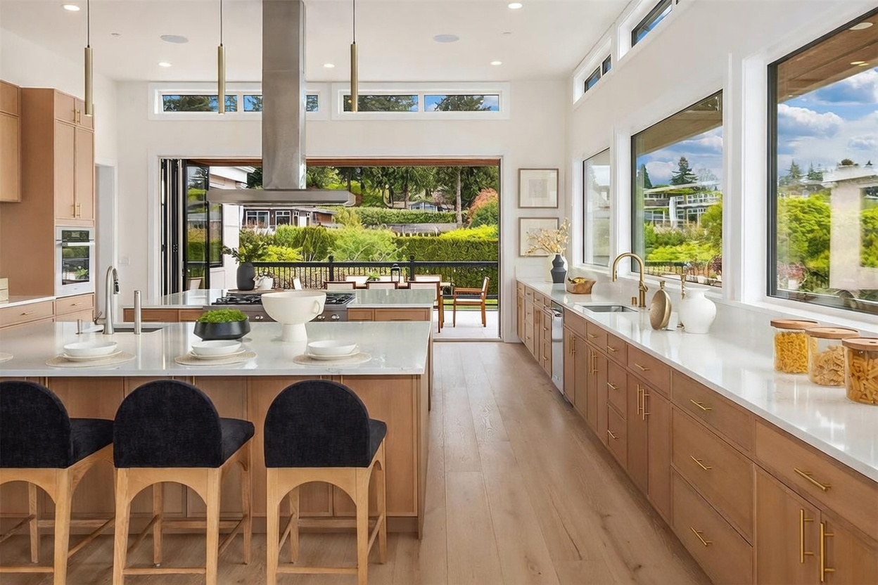 This bright and modern kitchen features light wood cabinetry, a large central island with seating, and expansive windows that provide a seamless transition to the outdoor deck. The space is defined by its clean lines, gold-toned hardware, and a professional-grade range hood, creating an airy and sophisticated atmosphere. The perspective looks directly through the open folding doors toward the lush greenery of the backyard, emphasizing the indoor-outdoor living design.