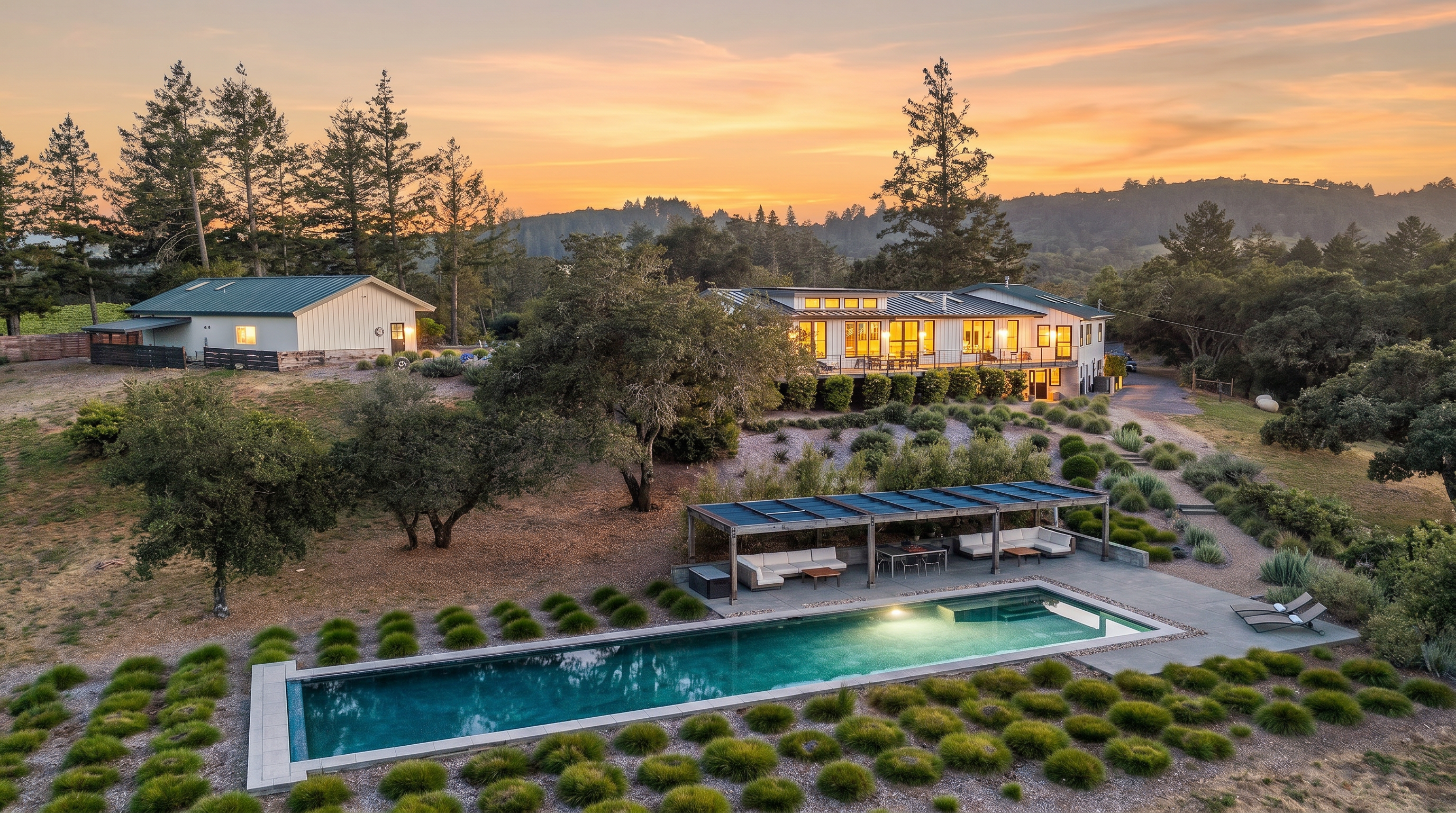 This high-angle aerial view captures a sprawling rural estate at dusk, featuring a modern farmhouse-style residence and a detached garage set against a backdrop of rolling hills and dense forest. In the foreground, a long, rectangular swimming pool is complemented by a shaded lounge area with outdoor seating, all surrounded by meticulously landscaped gardens with ornamental grasses. The scene conveys a sense of serene luxury and privacy, with the warm glow of the house lights contrasting against the cooling twilight sky.