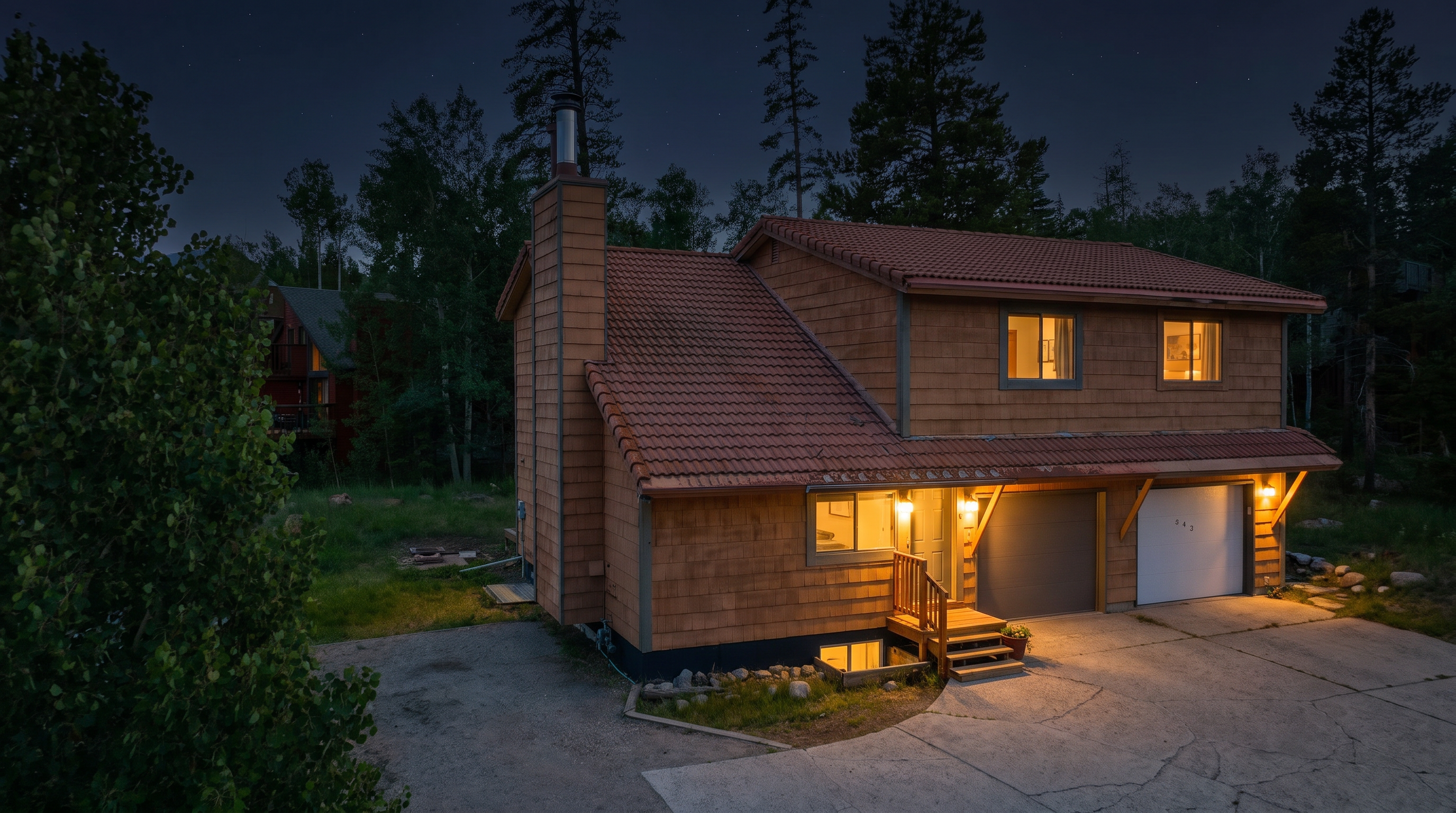 This two-story, wood-shingled residence is captured at twilight, showcasing its charming curb appeal and warm, inviting exterior lighting. The property features a two-car garage, a small wooden entry deck, and a classic tile roof, all nestled against a dense forest backdrop. A paved driveway leads to the home, creating an inviting, cozy atmosphere that highlights its functional yet picturesque design.