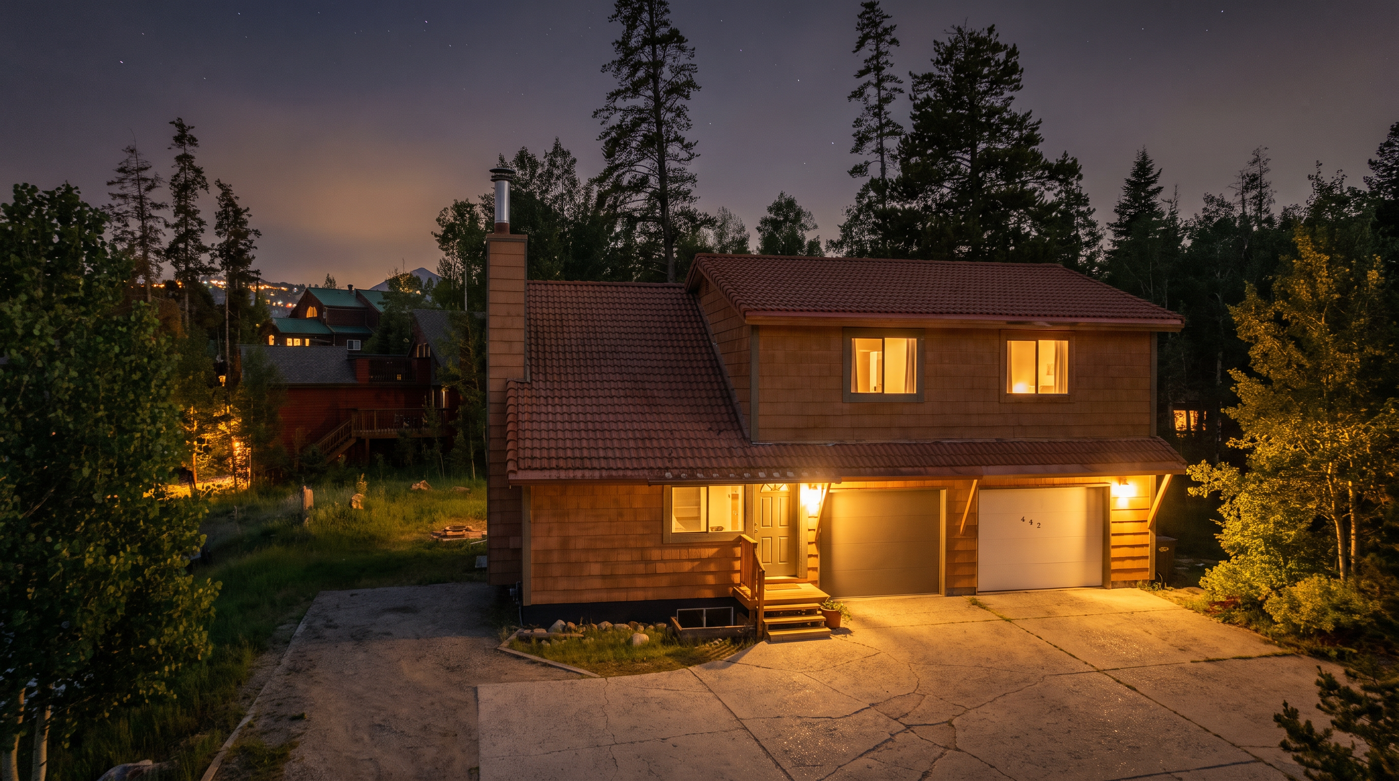 This two-story wooden home features a classic mountain-style design with a distinctive tiled roof and a two-car garage. The front facade is warmly illuminated, highlighting the entryway stairs, driveway, and large upper-story windows set against a backdrop of towering pine trees under a starry night sky. The property offers a cozy, inviting atmosphere, perfect for a peaceful woodland retreat.