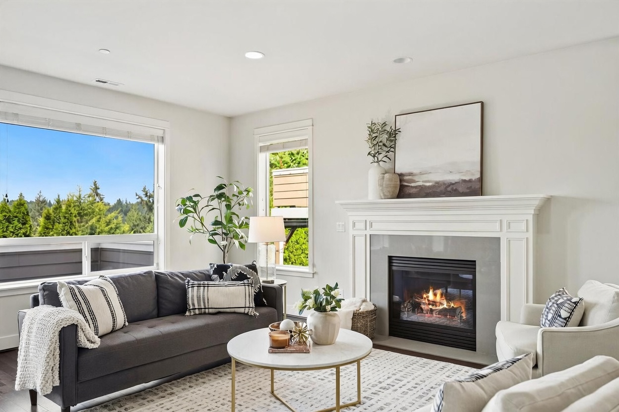 This bright and inviting living room features a dark gray sofa accented with patterned throw pillows and a cozy throw blanket. A central round coffee table with a light-colored top sits on a textured area rug, while a fireplace with a white mantel and a gray tiled surround serves as the room's focal point. Large windows provide ample natural light and a view of the surrounding trees, creating a warm and comfortable atmosphere.