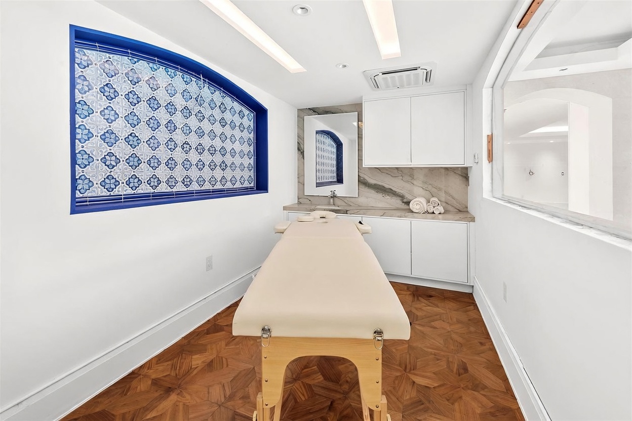 This specialized spa or massage room features a central treatment table set against a backdrop of white cabinetry and a stone-topped vanity. A striking arched window with blue and white patterned tile accents serves as a focal point, complemented by warm wood flooring in a herringbone pattern. The room is brightly lit with recessed ceiling lighting, creating a clean, professional, and serene atmosphere.