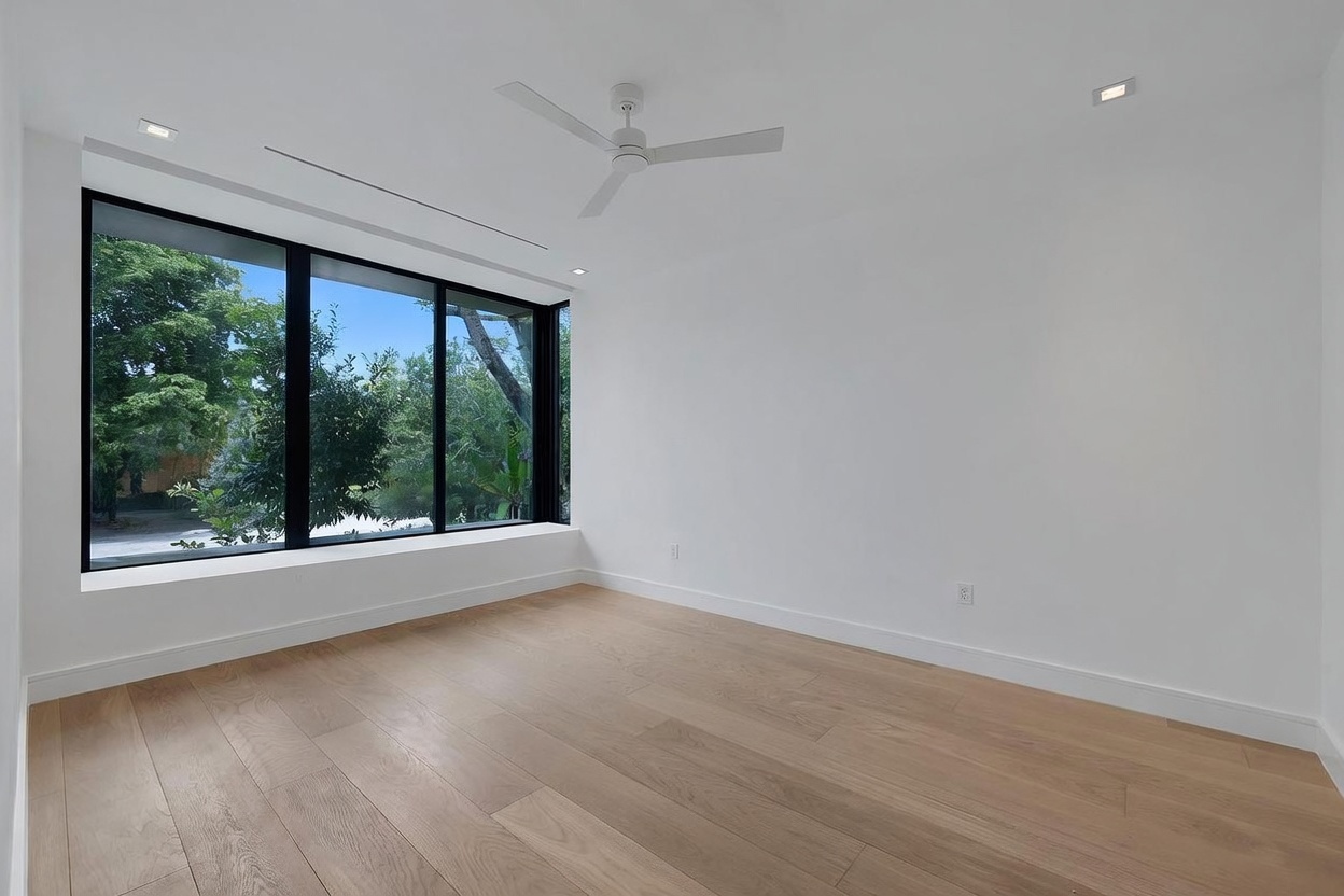 This minimalist, unfurnished room features light wood flooring and crisp white walls, creating a bright and airy atmosphere. A large window assembly provides an expansive view of the lush greenery outside, while a modern ceiling fan is centered above. The space is clean and neutral, offering a versatile canvas for any interior design style.