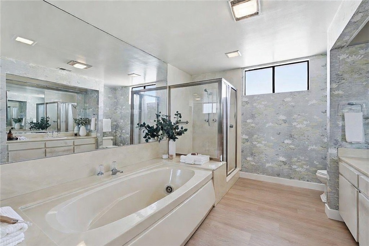 This spacious primary bathroom features a large soaking tub set against a mirrored wall, complemented by a separate glass-enclosed shower. The room is accented with patterned wallpaper and light-toned wood-look flooring, creating a bright and airy atmosphere. A double vanity with ample counter space and storage is visible, reflecting the clean and functional design of the space.