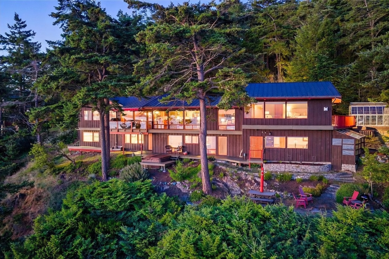 This elevated perspective captures a multi-level, wood-sided home nestled into a lush, forested hillside. The residence features expansive decks, large windows that illuminate the interior, and a private hot tub overlooking the landscape, all set against a backdrop of dense trees. A stone patio area with a fire pit and outdoor seating sits at the base of the home, creating a serene and secluded atmosphere.