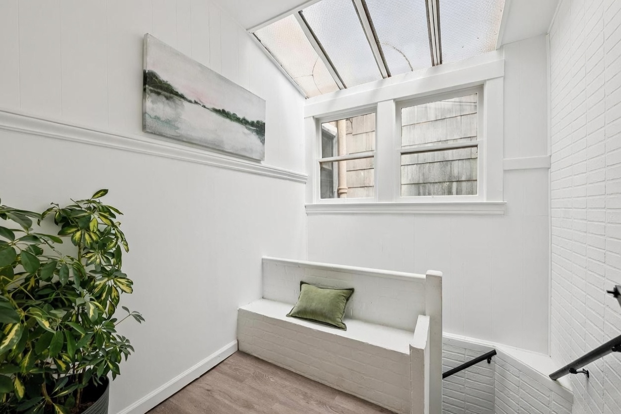 This bright, transitional space features a built-in white brick bench topped with a green accent pillow, positioned next to a staircase leading downward. A large skylight overhead floods the area with natural light, highlighting the clean white walls and light wood-toned flooring. A piece of landscape art hangs above the bench, and a potted plant adds a touch of greenery to the corner, creating a serene and inviting atmosphere.
