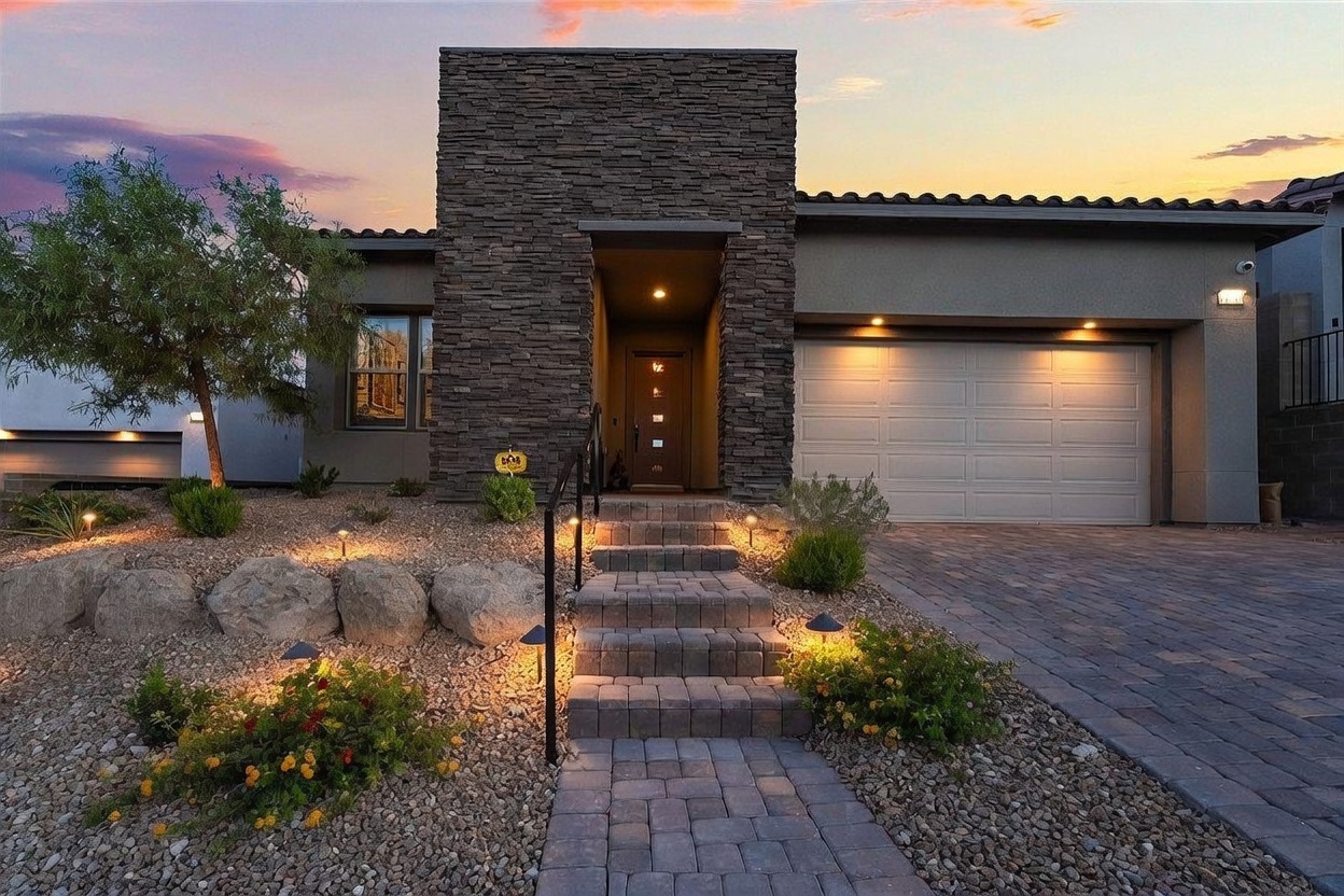 This modern single-story home features a striking facade with a prominent, stacked-stone entryway tower that creates a bold architectural focal point. A paved walkway with stone steps leads up to the front door, flanked by low-maintenance desert landscaping and integrated landscape lighting. The exterior is finished in a neutral tone, complemented by a multi-car garage and a warm, inviting glow from the evening sky.