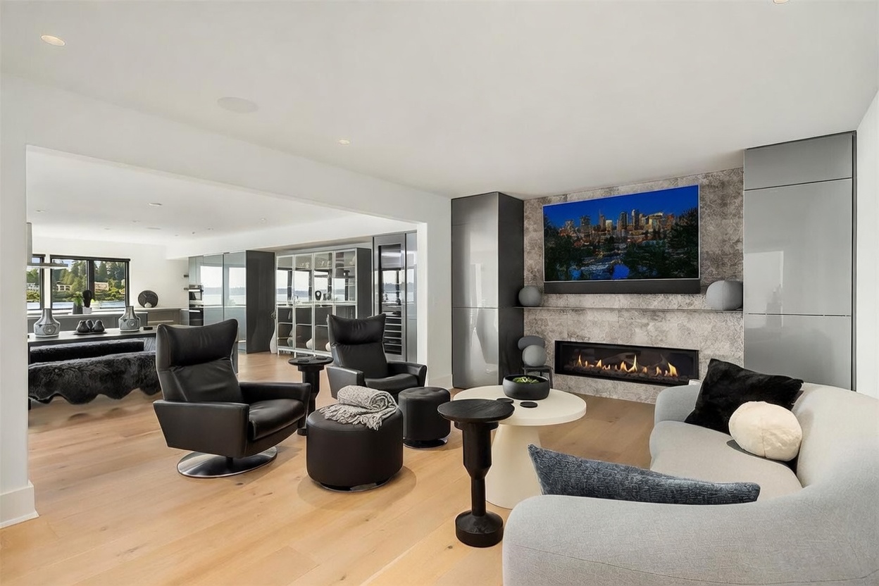 This contemporary open-concept living space features light wood flooring and a sophisticated neutral color palette. A sleek linear fireplace is set into a textured stone-clad wall, topped by a large wall-mounted television, while comfortable leather armchairs and a curved sofa create an inviting seating area. The room flows seamlessly into an adjacent dining area, with large windows providing a view of the water beyond.
