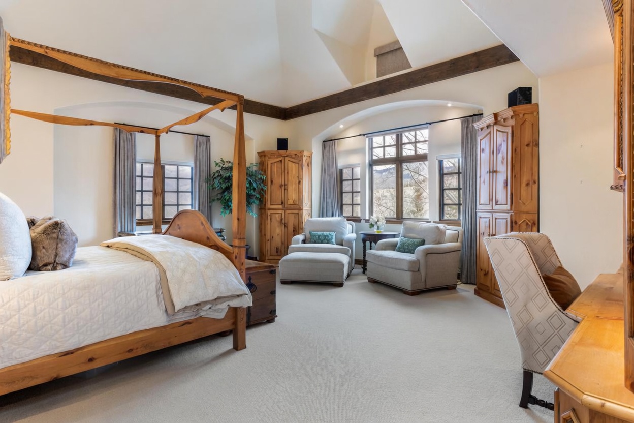This spacious primary bedroom features a prominent four-poster wooden bed frame and a cozy seating area with two armchairs and an ottoman positioned near a large window arrangement. The room is accented by exposed wooden ceiling beams, light-toned carpeting, and two tall wooden armoires that provide ample storage. The perspective captures the entire suite, highlighting the blend of rustic wood elements and comfortable, neutral-toned furnishings.