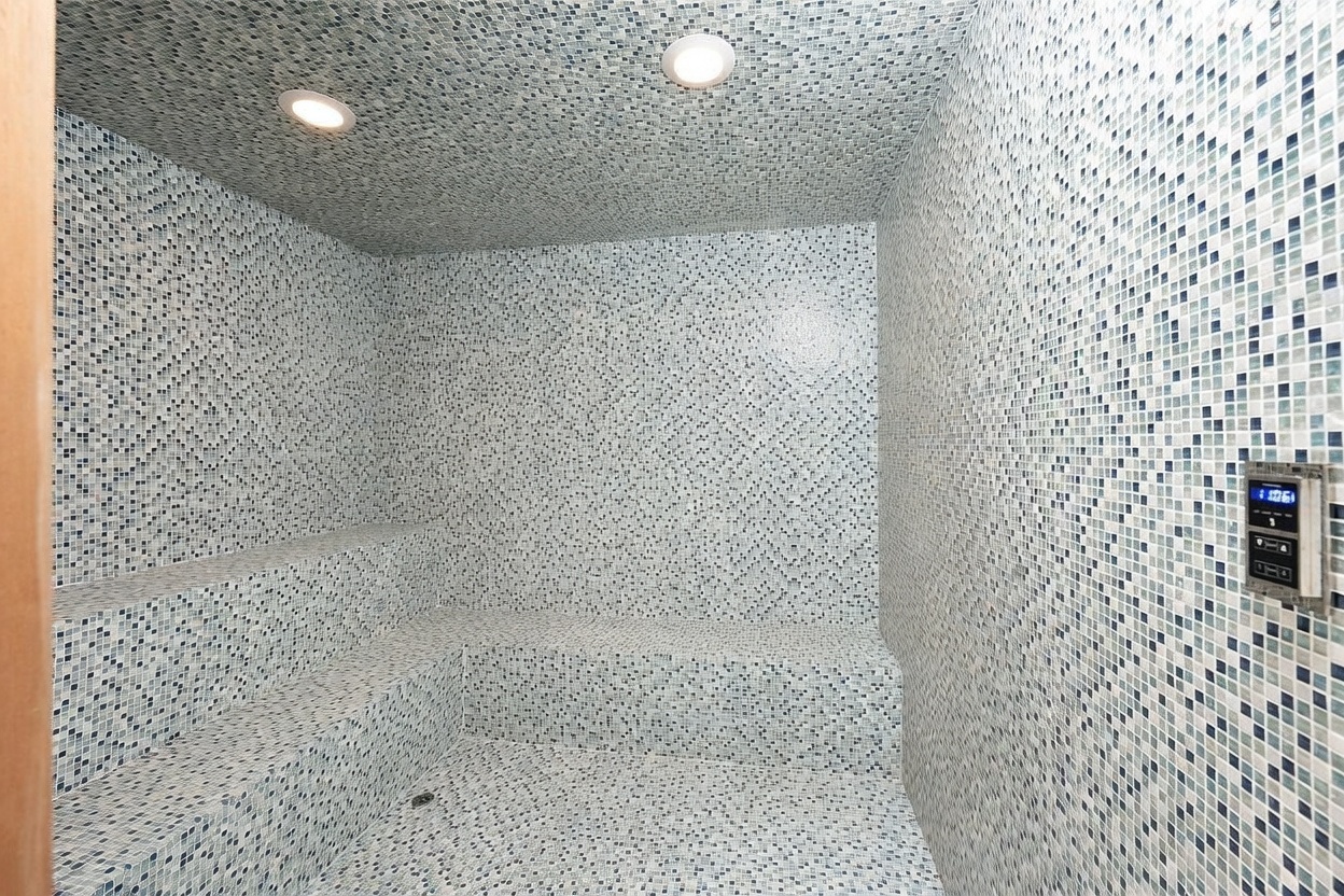 This image features a steam room or sauna completely finished in small, light-toned mosaic tiles. The space includes built-in tiled bench seating along the walls and is illuminated by two recessed ceiling lights. A digital control panel is mounted on the right wall to manage the room's temperature and settings.