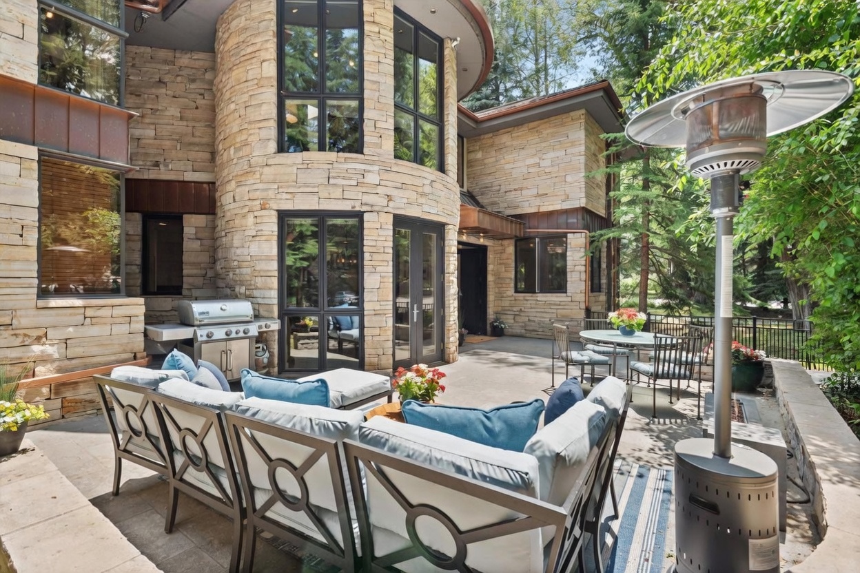 This spacious stone-paved patio features a comfortable outdoor seating area with plush cushions and a modern grill station. The home's exterior is characterized by curved stone walls and tall windows that create a sophisticated, architectural aesthetic. A tall outdoor heater and a small dining set complete the space, which is nestled against a lush, wooded backdrop.