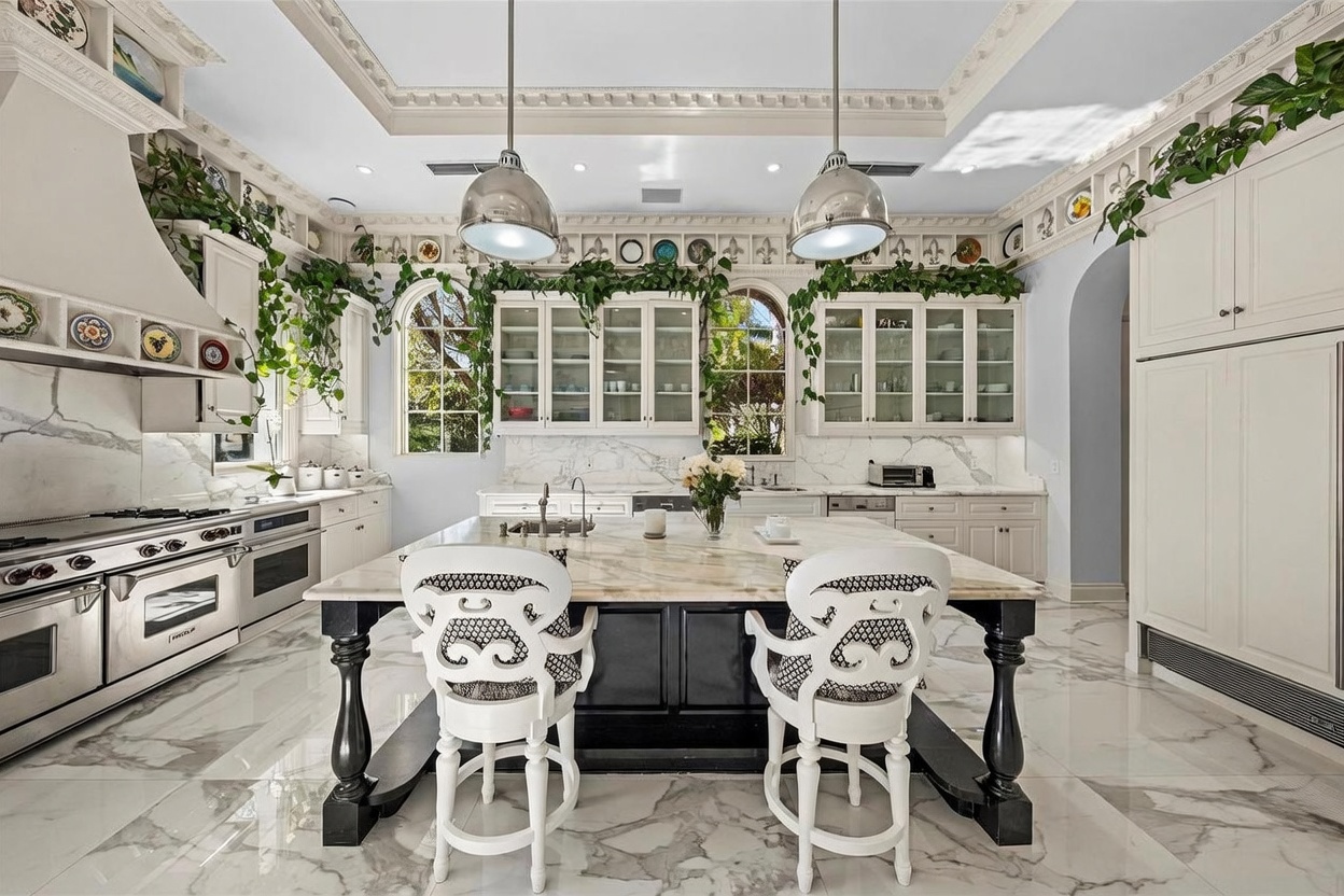This elegant kitchen features a large central island with a stone countertop and a dark base, complemented by two ornate white chairs. The space is defined by white cabinetry, stainless steel appliances, and decorative plates displayed along the upper wall molding. Large arched windows allow natural light to fill the room, while trailing greenery adds a vibrant, organic touch to the sophisticated, classic design.