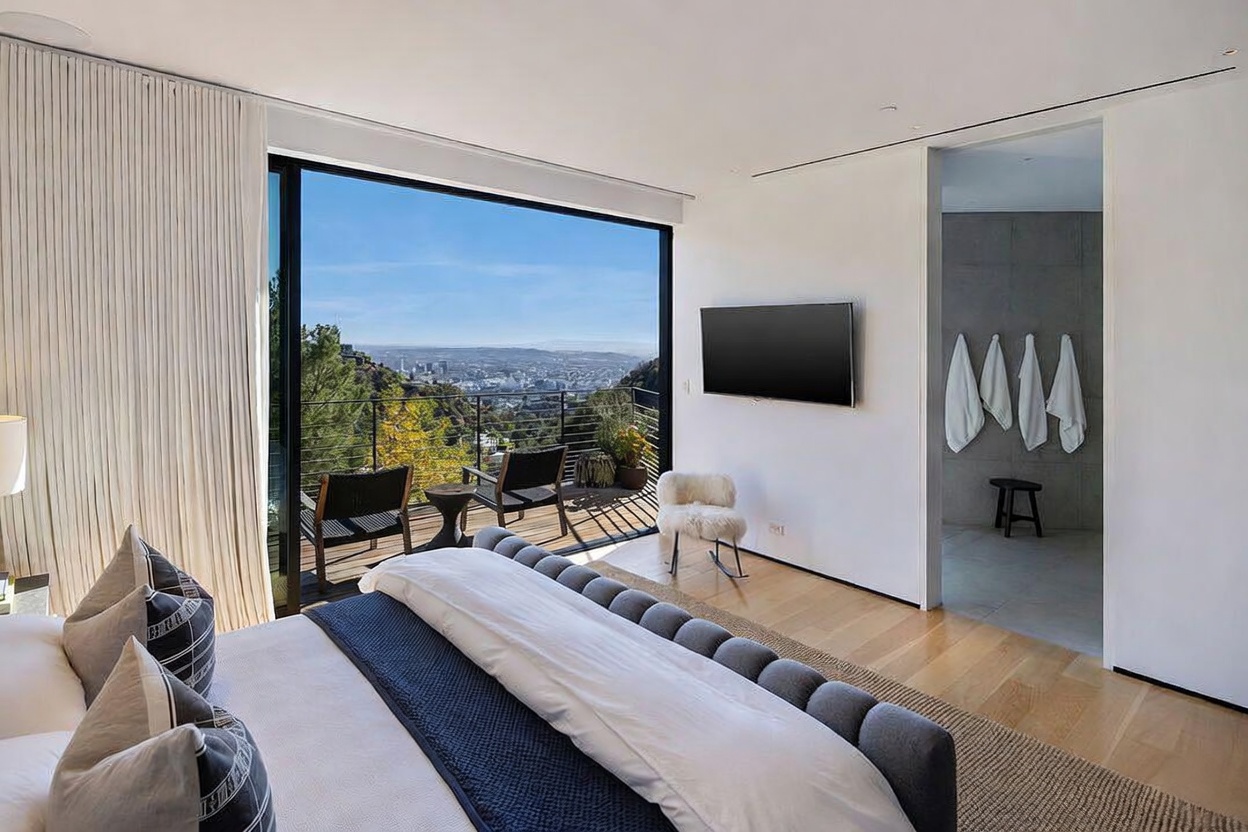 This primary bedroom features a contemporary design with a large bed as the focal point, positioned to take advantage of the expansive view through floor-to-ceiling glass doors. The room opens onto a private deck with outdoor seating, overlooking a scenic hillside landscape and city skyline. A doorway leads into an adjacent bathroom, which is equipped with hanging towels and a small stool, maintaining a minimalist and serene aesthetic.