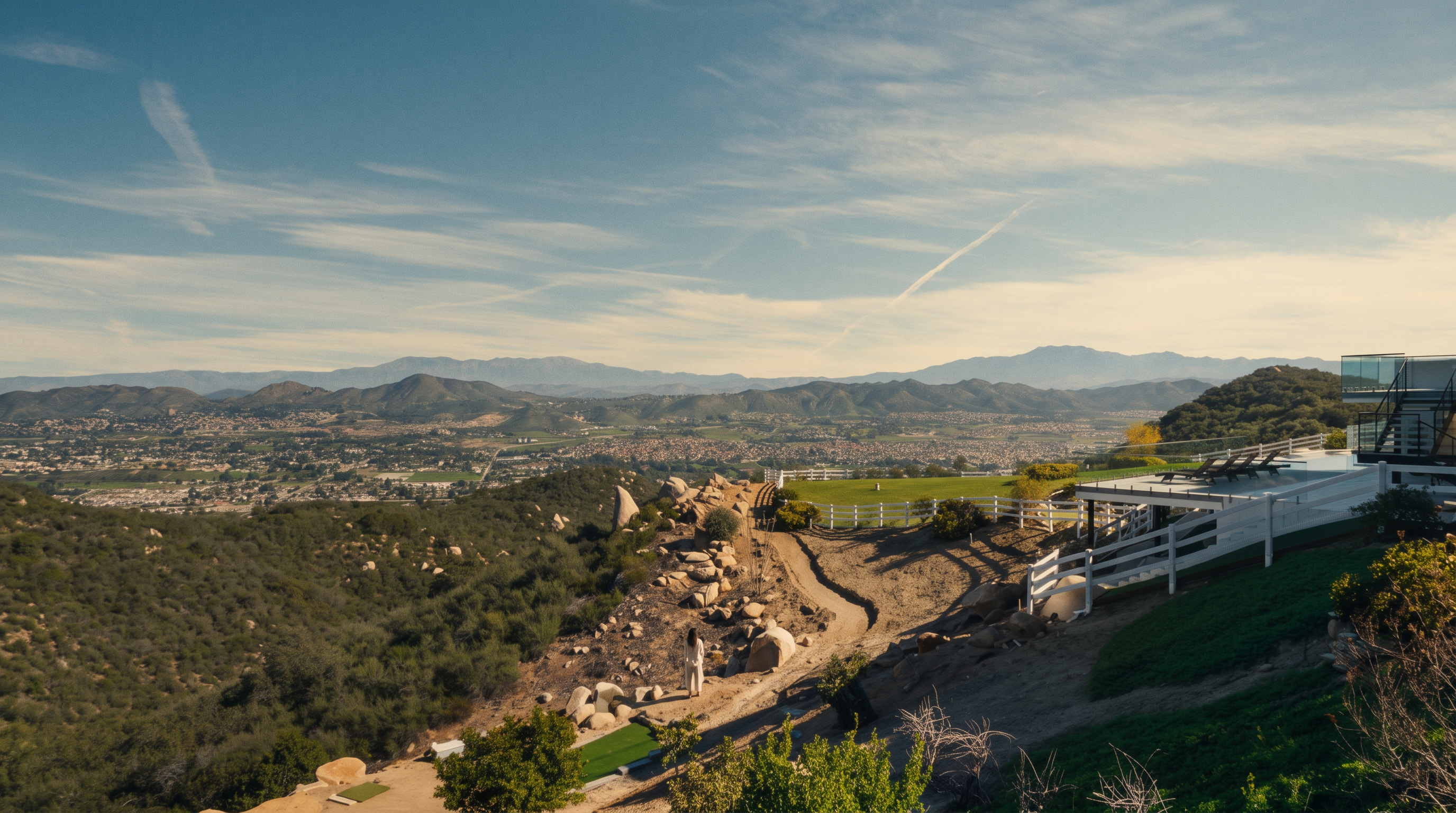 This panoramic rear exterior shot highlights a spacious hillside property with expansive views of the valley and mountains. The home features a multi-level deck and patio area designed for outdoor living, complemented by a white fence and lush green landscaping. Its elevated position offers a sense of privacy and luxury, perfect for scenic entertaining.