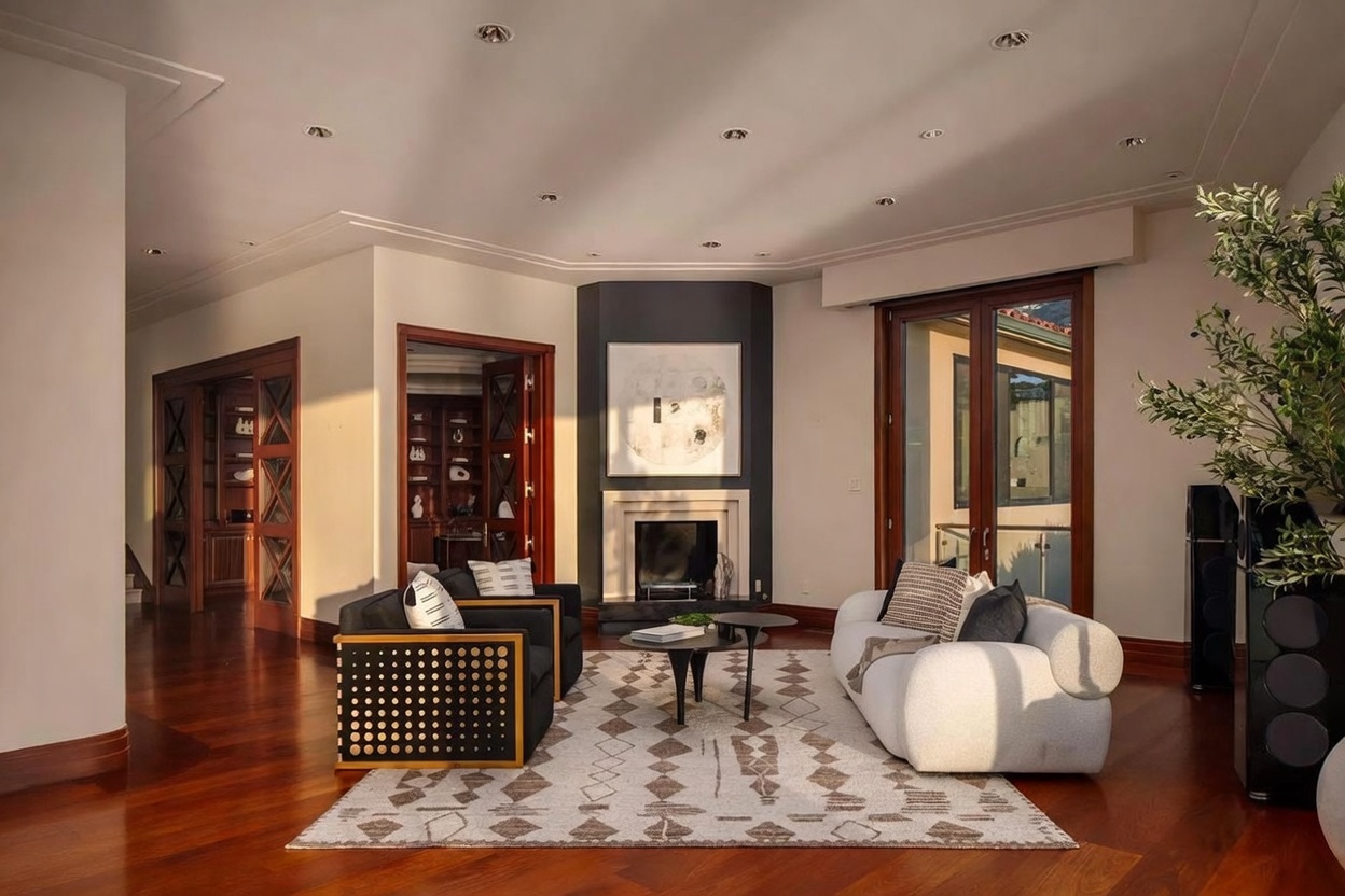 This sophisticated living room features rich hardwood flooring and a central fireplace with a dark, dramatic surround and a large piece of abstract art above. The space is furnished with a modern white curved sofa and two dark armchairs arranged around a pair of circular coffee tables on a patterned area rug. Large glass doors provide access to an outdoor space, allowing natural light to fill the room, while recessed lighting adds a warm, inviting glow to the interior.