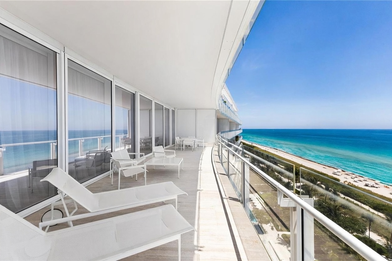 This expansive, curved balcony offers a luxurious outdoor living space with panoramic views of the turquoise ocean and sandy coastline. The area is furnished with white lounge chairs and a dining set, all set against a backdrop of floor-to-ceiling glass walls that seamlessly connect the interior to the exterior. The sleek, modern design emphasizes a bright, airy atmosphere perfect for coastal relaxation.
