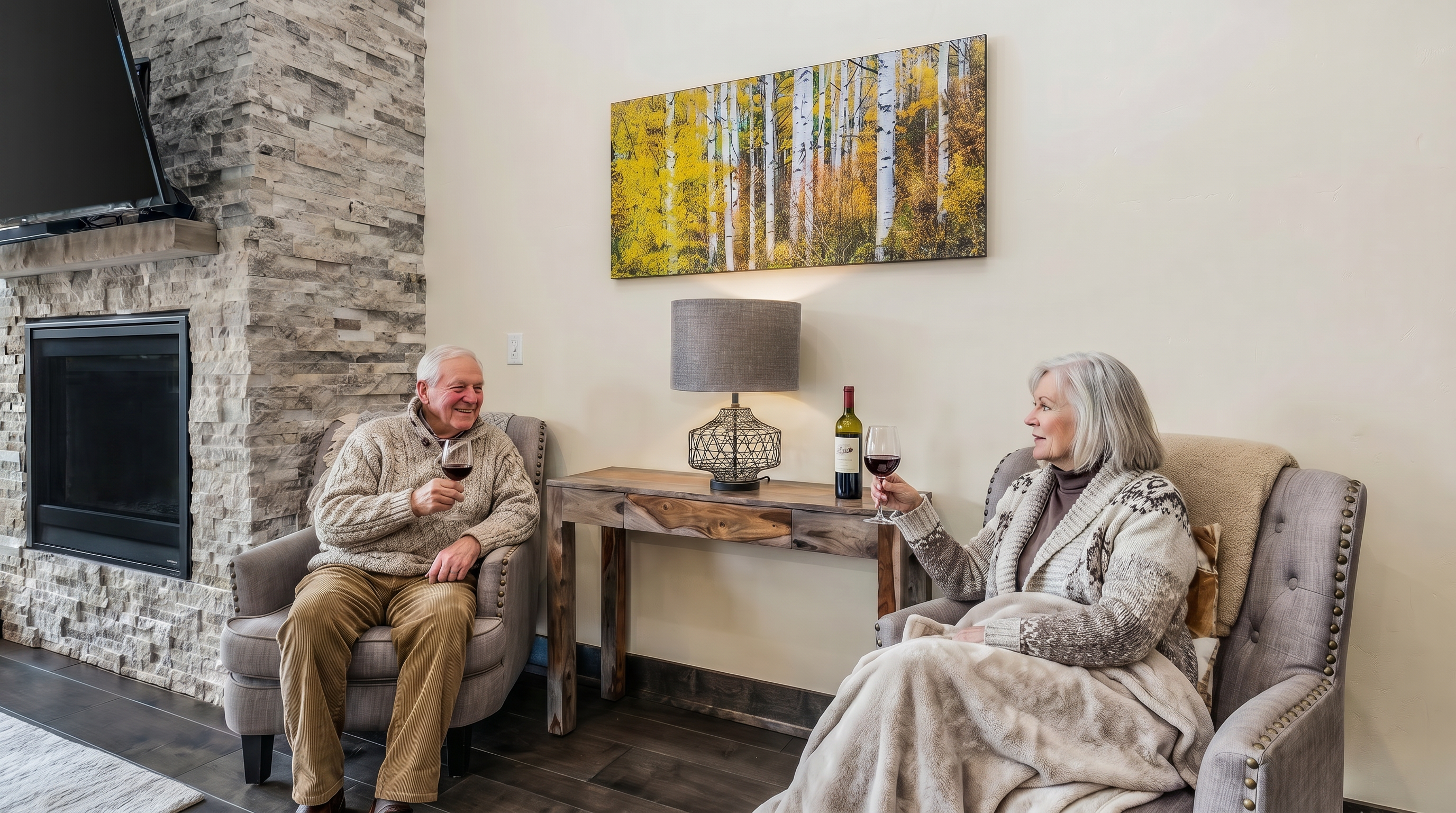 This cozy living room area features a stone-clad fireplace with a mounted television, flanked by two comfortable grey upholstered armchairs. A rustic wooden console table sits between the chairs, topped with a decorative wire-frame lamp and positioned beneath a vibrant landscape painting of an aspen forest. The warm lighting and natural textures create an inviting, rustic-chic atmosphere perfect for relaxation.