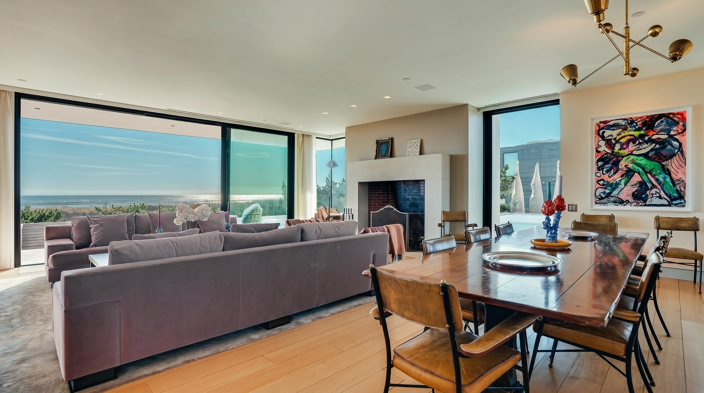 This open-concept living and dining area features a large, plush purple sectional sofa facing a stone-clad chimney breast with a brick firebox. To the right, a long wooden dining table is surrounded by leather-upholstered chairs, illuminated by a modern multi-arm chandelier. The space is defined by light wood flooring and expansive glass walls that provide panoramic views of the ocean and the outdoor pool area.