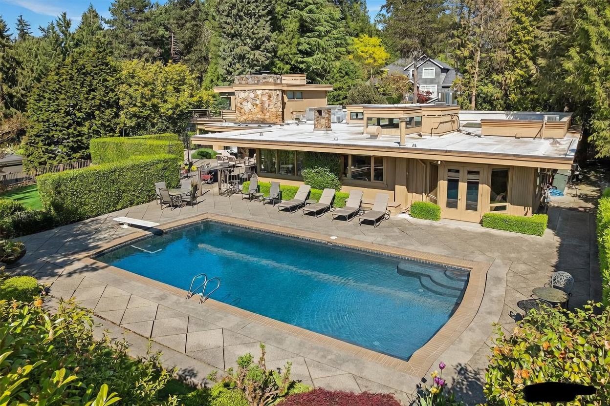 This elevated perspective captures a sprawling backyard featuring a large, rectangular swimming pool with a diving board and integrated steps. The surrounding concrete patio is furnished with multiple lounge chairs and a dining set, all set against a backdrop of lush, mature trees and manicured hedges. The home itself showcases a mid-century modern aesthetic with a flat roofline and a prominent stone-clad chimney breast.