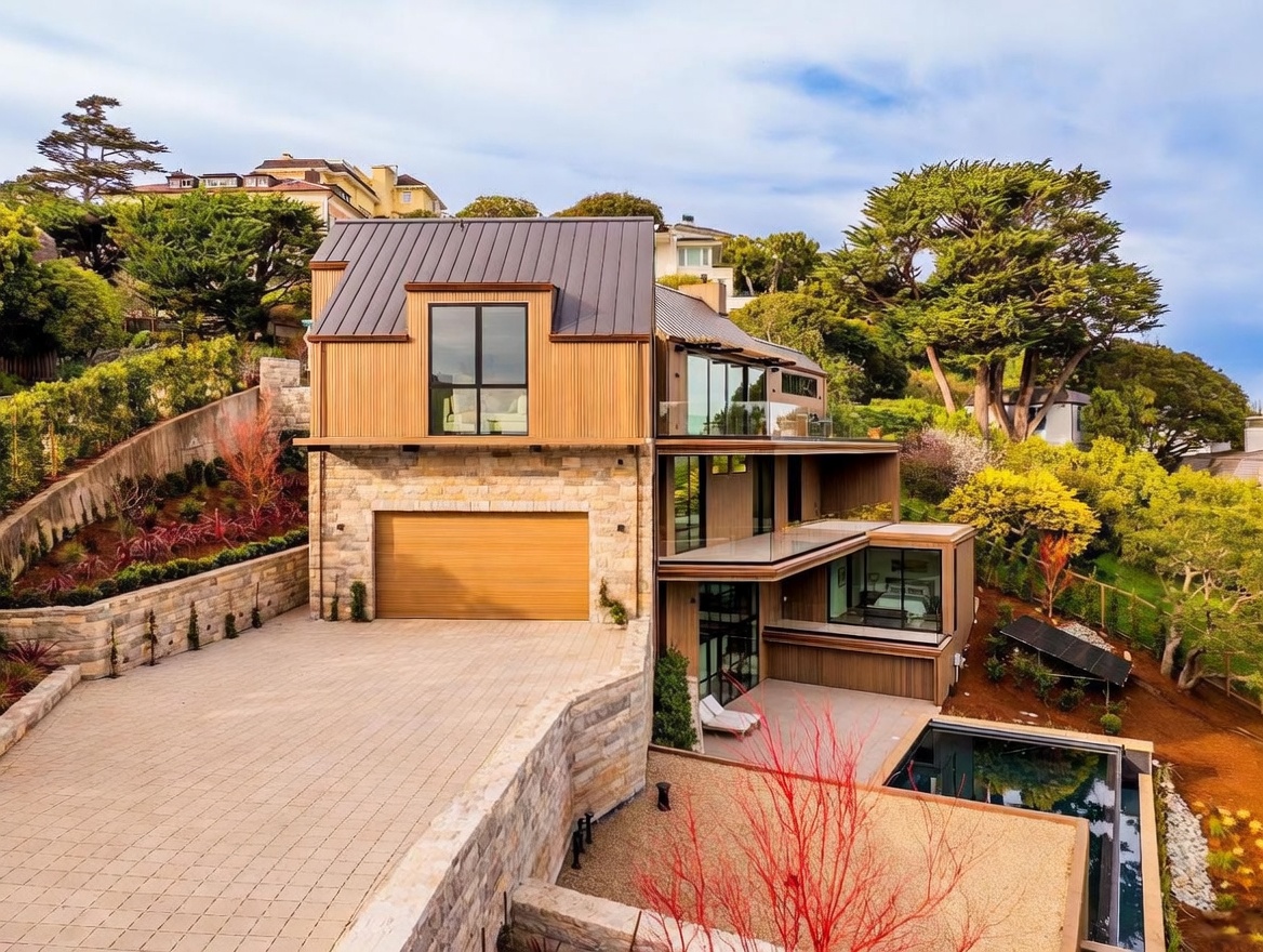 This modern multi-level residence features a striking combination of natural stone masonry on the lower levels and vertical wood siding on the upper structure. The home includes a spacious paved driveway leading to a garage, multiple levels of balconies with glass railings, and a private swimming pool set within a landscaped yard. The architectural design emphasizes clean lines and large windows, creating a sophisticated and contemporary aesthetic set against a backdrop of mature trees.