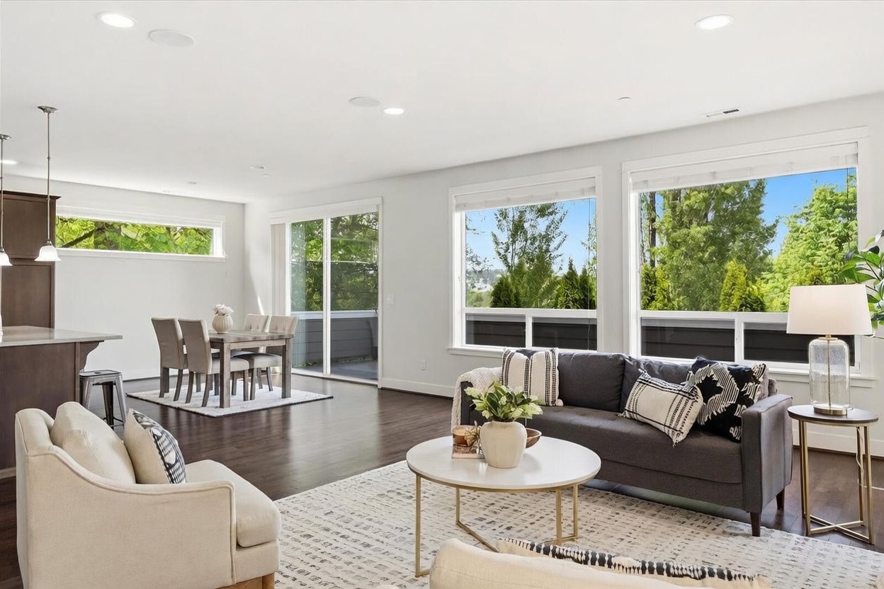 This bright and open-concept living area features dark hardwood flooring and a neutral color palette, creating a sophisticated and airy atmosphere. The space is anchored by a dark gray sofa and a round coffee table, while large windows provide abundant natural light and views of the surrounding greenery. A dining area is visible in the background, seamlessly integrated into the main living space, making it ideal for modern entertaining.