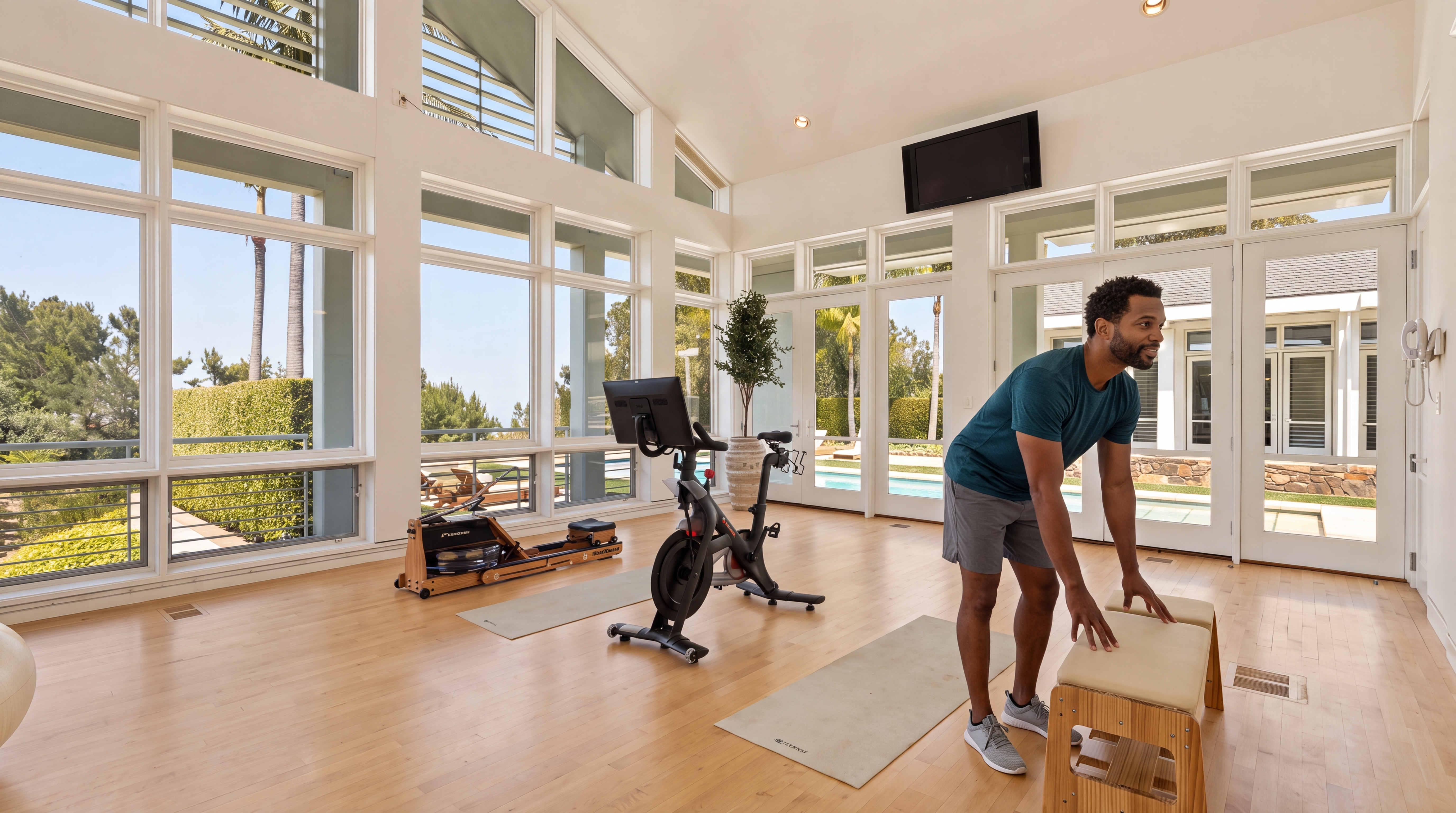 This bright and airy home gym features vaulted ceilings and floor-to-ceiling windows that provide abundant natural light and scenic views of the outdoor landscape. The space is equipped with a stationary bicycle, a rowing machine, and pilates equipment, offering a functional and motivating workout environment. Its open-concept design with light-toned hardwood flooring enhances the room's spacious and modern aesthetic.