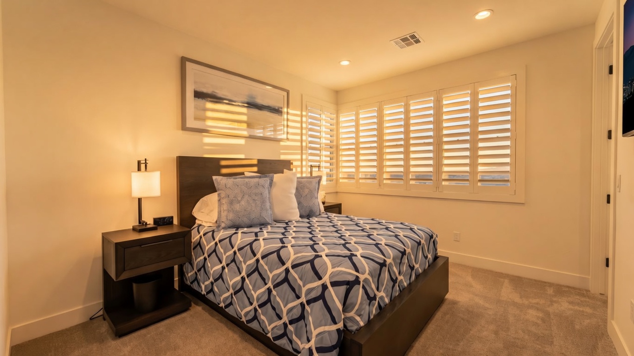 This well-lit bedroom features a dark wood bed frame centered against a white wall, accented by a large piece of abstract wall art. The room is furnished with a matching dark wood nightstand and a lamp, while white plantation shutters cover the windows to provide adjustable natural light. The space is finished with neutral carpeting, creating a clean and inviting atmosphere.