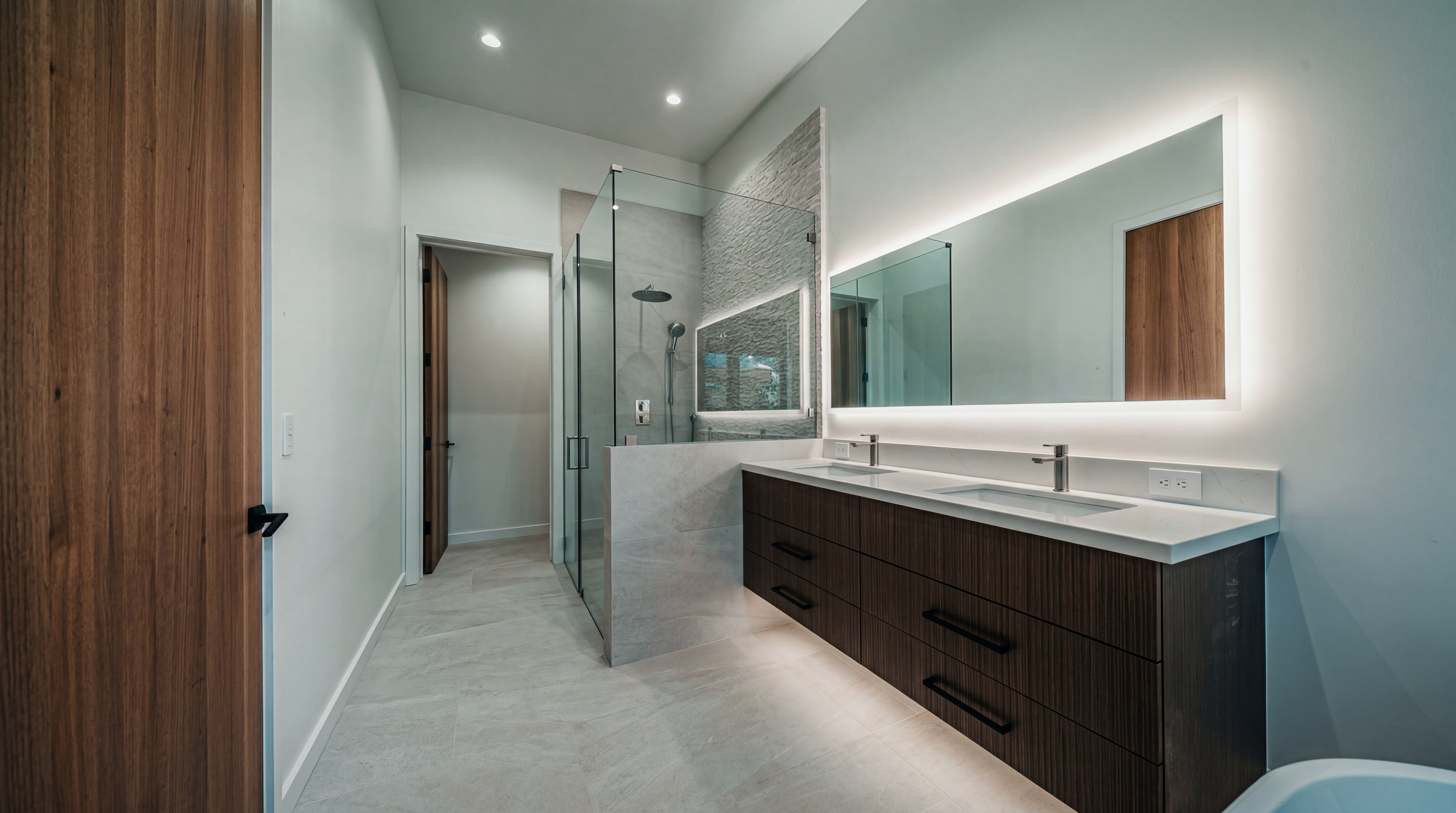 This modern primary bathroom features a floating double vanity with dark wood-toned cabinetry and a white countertop. A large, illuminated mirror hangs above the vanity, while a glass-enclosed walk-in shower with textured stone wall accents sits to the left. The space is finished with light-colored tile flooring and minimalist fixtures, creating a clean, spa-like atmosphere.