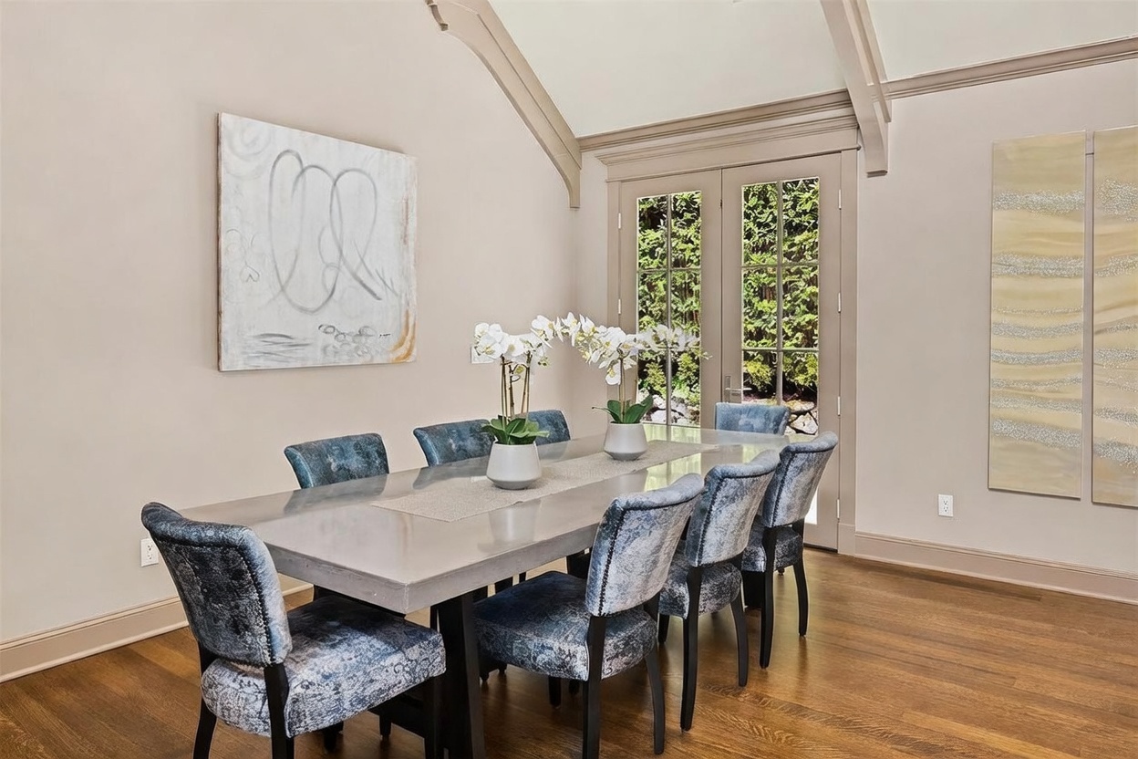 This elegant dining room features a long, smooth-topped table surrounded by eight upholstered chairs in a textured blue fabric. The space is highlighted by a vaulted ceiling with decorative beams and tall glass doors that provide a view of the lush greenery outside. Two large pieces of abstract wall art and a pair of white orchid centerpieces add a sophisticated, curated touch to the neutral-toned room.