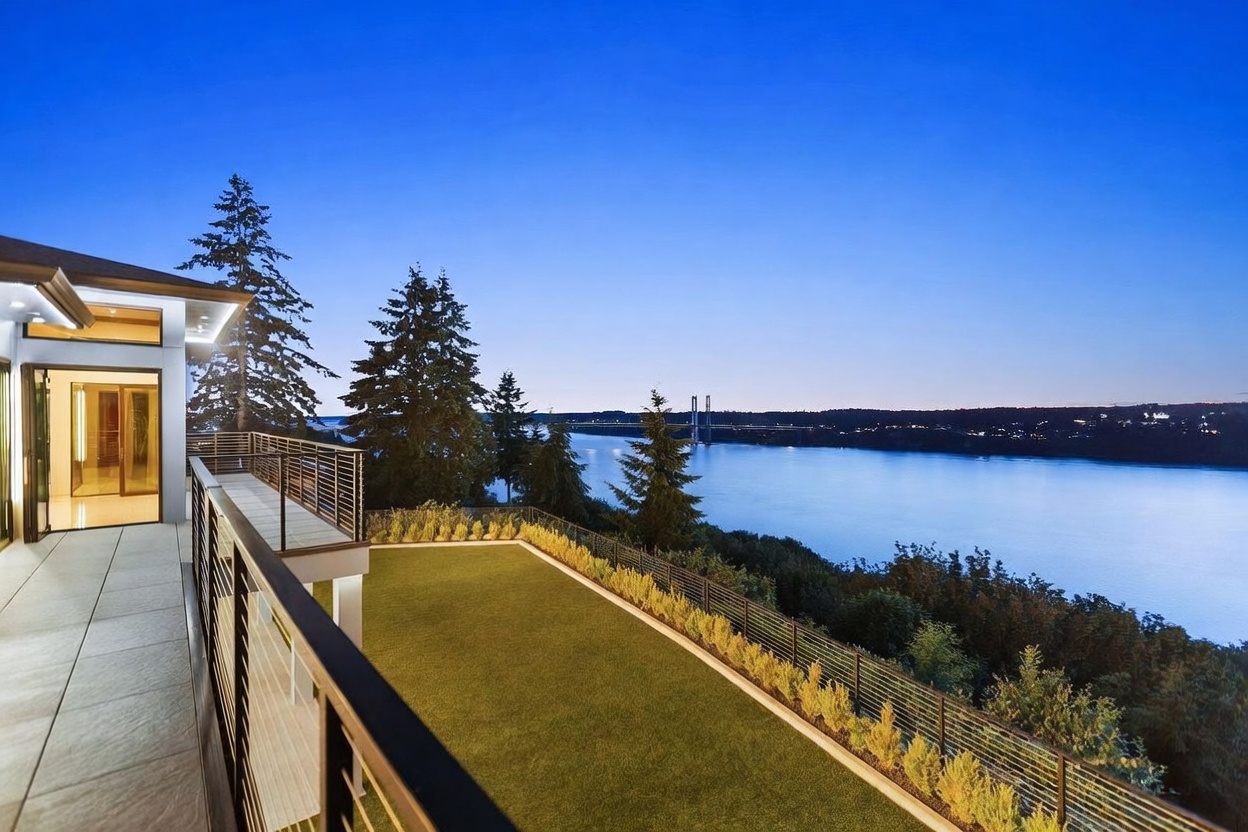 This elevated balcony offers a panoramic view of a wide river and a distant bridge under a clear twilight sky. The space features a sleek metal railing, light-colored tiled flooring, and a manicured lawn area that overlooks the natural landscape below. The modern architectural design emphasizes indoor-outdoor living, with large glass doors providing seamless access to the home's interior.