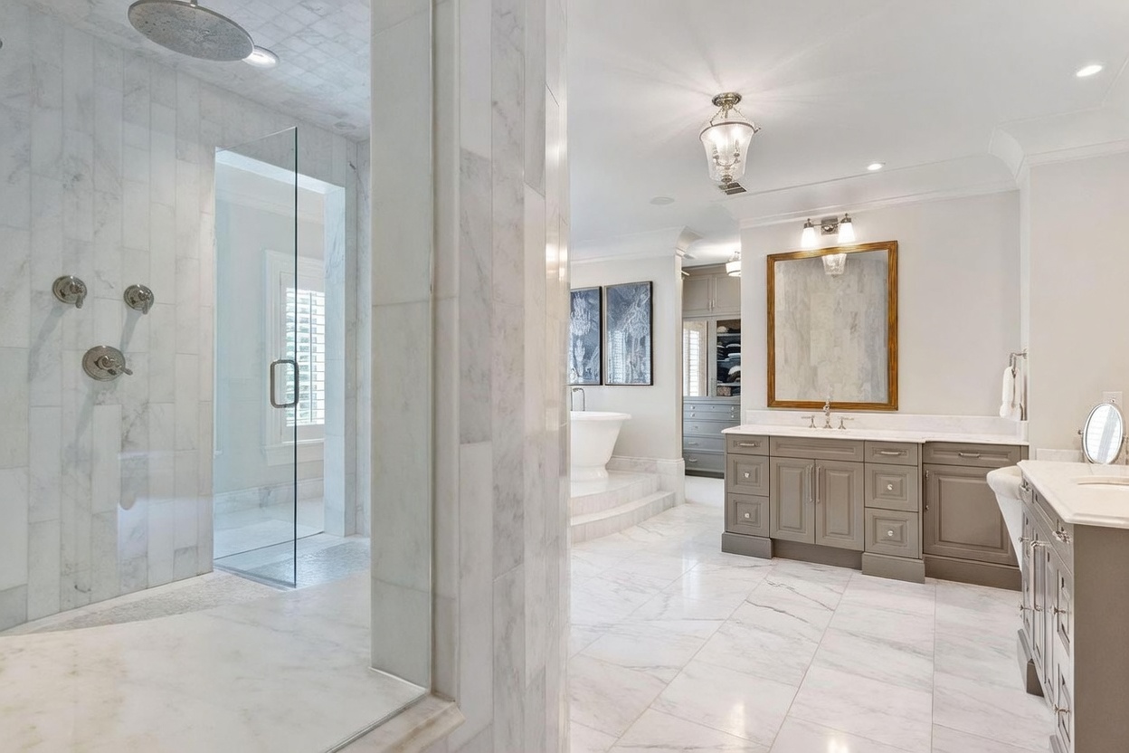 This luxurious primary bathroom features a spacious walk-in shower with glass panels and a rainfall showerhead, adjacent to a vanity area with a gold-framed mirror and gray cabinetry. A freestanding soaking tub is positioned on a raised platform in the background, creating a spa-like atmosphere. The room is finished with light-toned stone flooring and walls, illuminated by a decorative chandelier and recessed lighting.