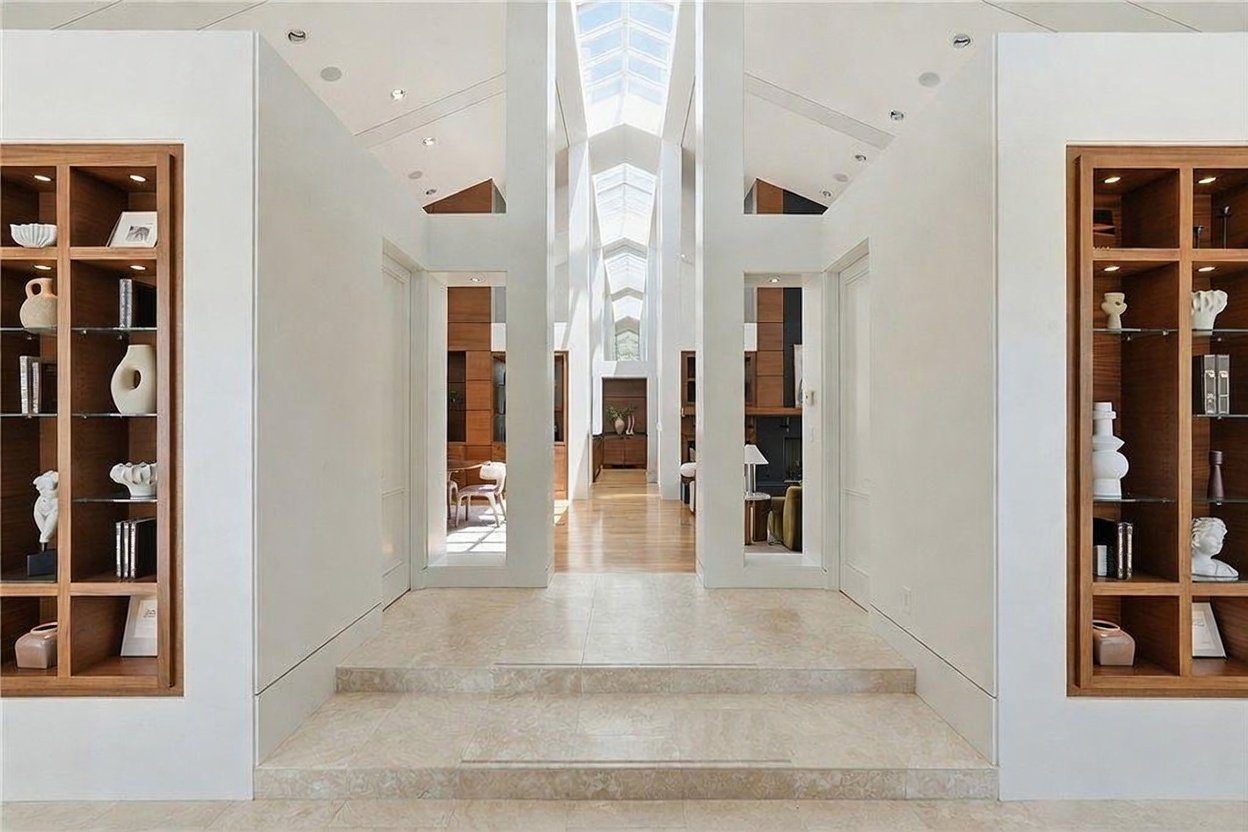 This grand, symmetrical entryway features a central stone-tiled staircase leading into a bright, airy hallway with a dramatic skylight running the length of the ceiling. Flanking the central path are built-in wooden shelving units displaying decorative objects, creating a sophisticated and balanced architectural aesthetic. The perspective is centered and cinematic, drawing the eye through the open-plan layout toward the distant living spaces.