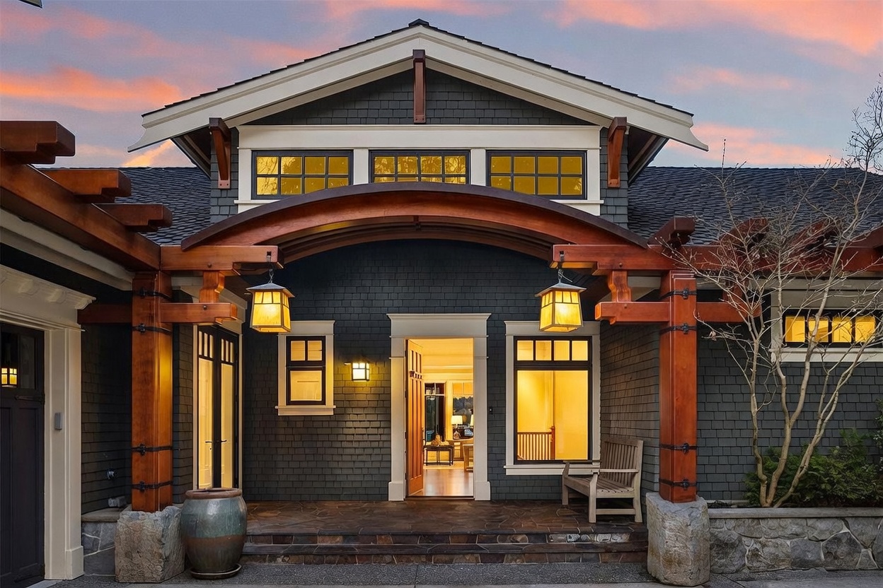 This stunning Craftsman-style entryway features a dramatic arched timber pergola supported by stone pillars, creating a warm and inviting focal point. The dark shingle siding contrasts beautifully with the rich wood tones and glowing lantern-style lighting, while the open front door offers a glimpse into the bright, welcoming interior. The scene is captured at dusk, with a soft sunset sky providing a picturesque backdrop for this elegant home entrance.