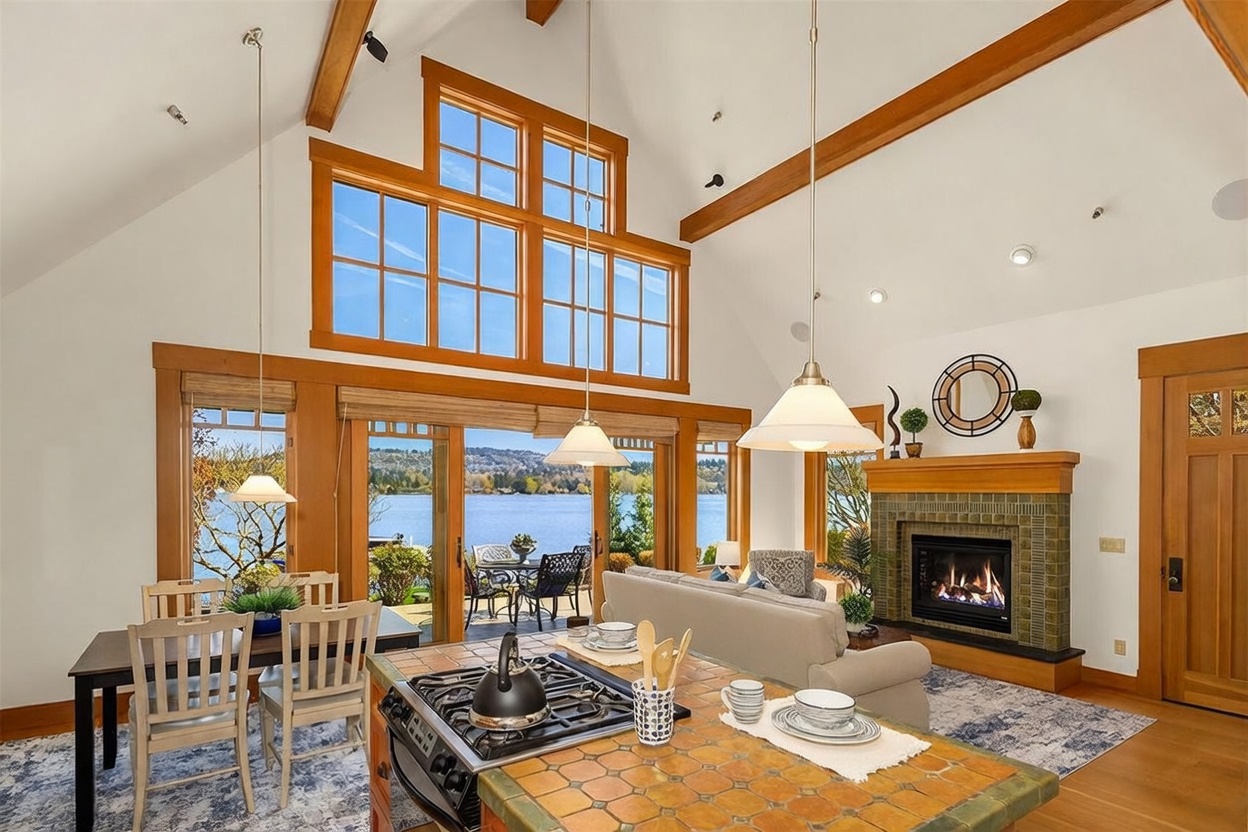 This open-concept living space features a kitchen island with a tiled countertop and a built-in gas cooktop, seamlessly flowing into a dining area and a cozy living room with a fireplace. The room is defined by its soaring vaulted ceilings with exposed wooden beams and a massive wall of windows that frame a stunning, direct view of the lake. The warm wood tones and natural light create an inviting, airy atmosphere perfect for waterfront living.