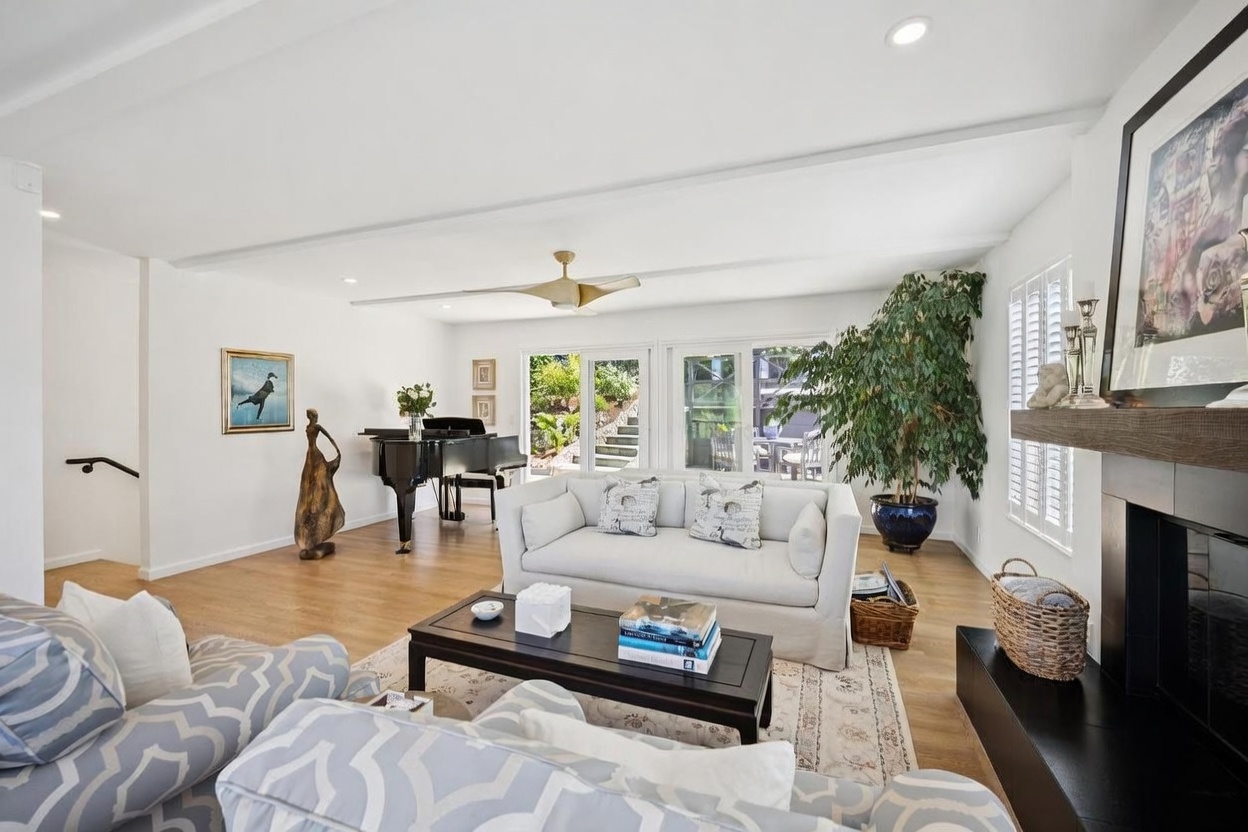 This bright and airy living room features light wood flooring and a neutral color palette, creating a welcoming and sophisticated atmosphere. The space is anchored by a comfortable sofa and a dark wood coffee table, with a grand piano positioned near large sliding glass doors that provide a view of the outdoor greenery. A tall fireplace surround with a wooden mantel adds a focal point to the room, while a large potted plant and curated artwork enhance the refined, open-concept design.